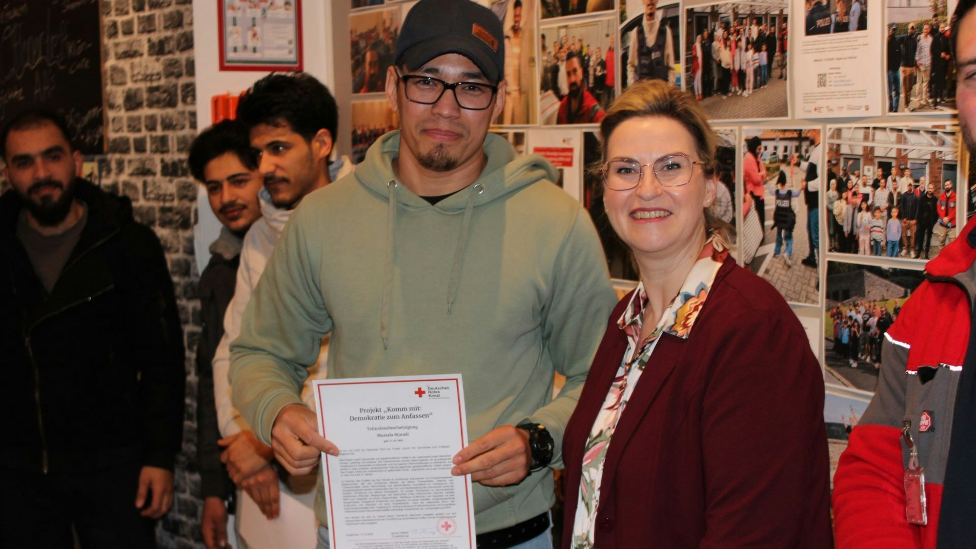 Der junge Afghane steht mit Sandra Eisermann vor einer Fotowand. In den Händen hält er sein Zertifikat.