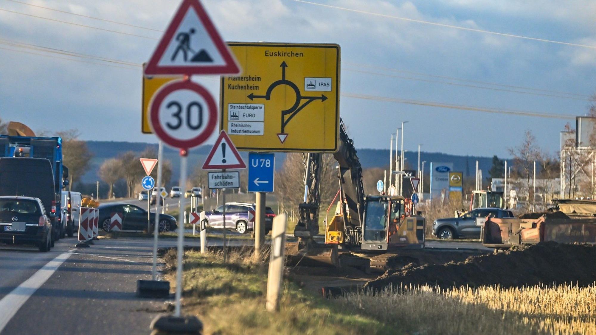 Schilder weisen auf eine Baustelle in Euskirche hin, ein Bagger steht neben einer stark befahrenen Straße.