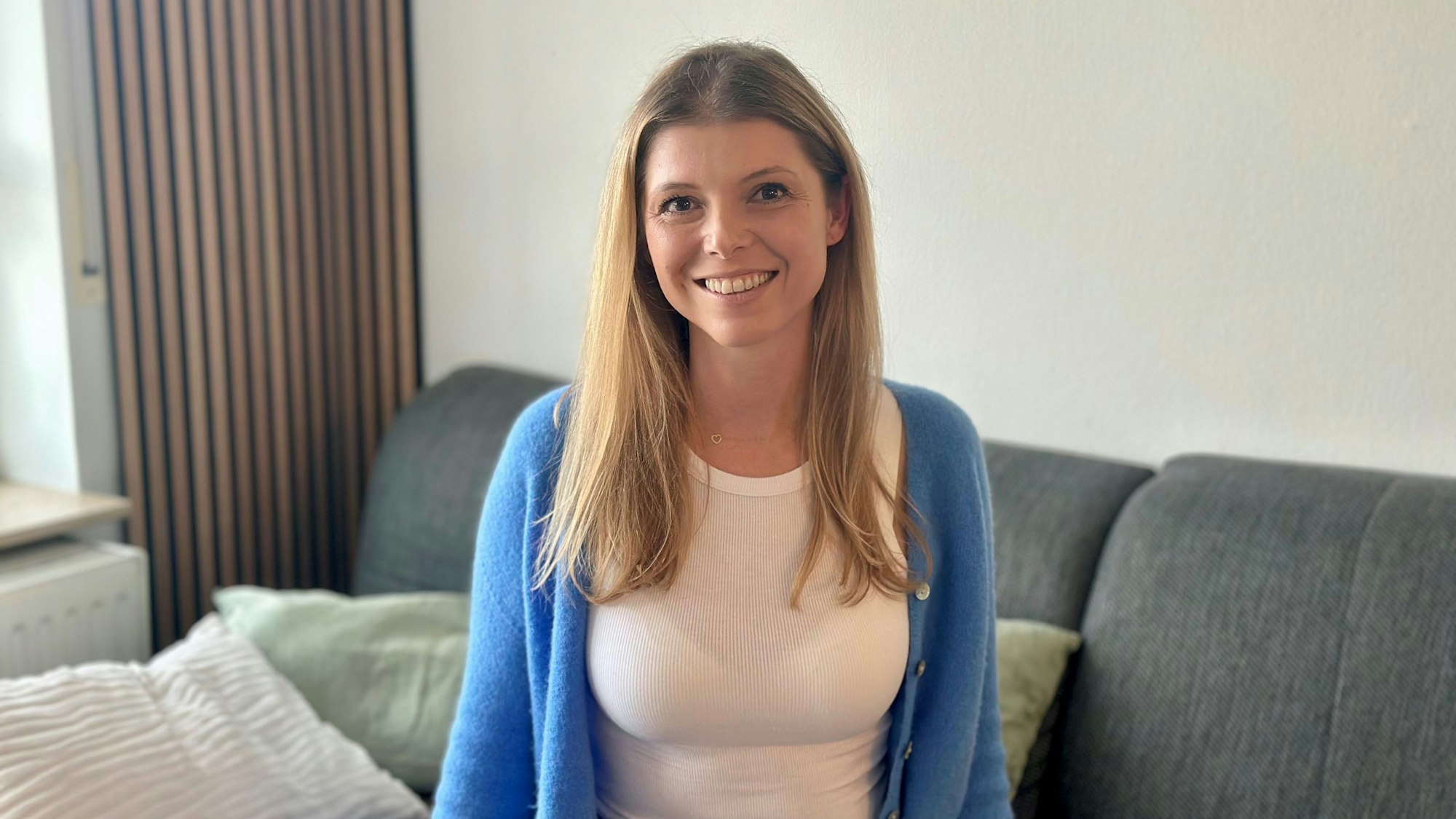 Eine blonde Frau in blauer Jacke sitzt auf einem grauen Sofa.
