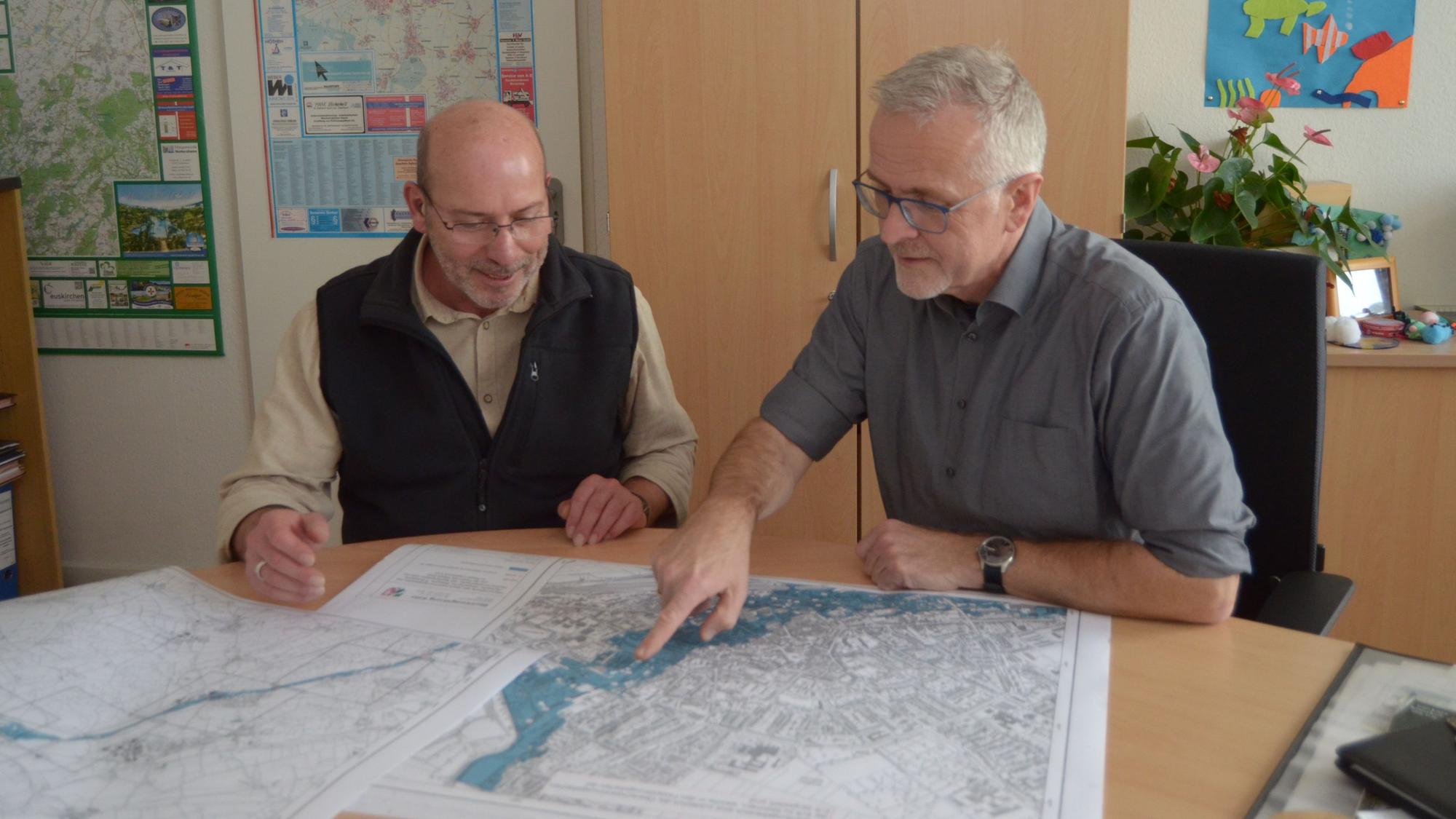 Bernd Kuballa (r.) und Norbert Wiedemann sitzen an einem Tisch. Wiedemann zeigt Kuballa einen Fluss auf einer ausgebreiteten Stadtkarte.