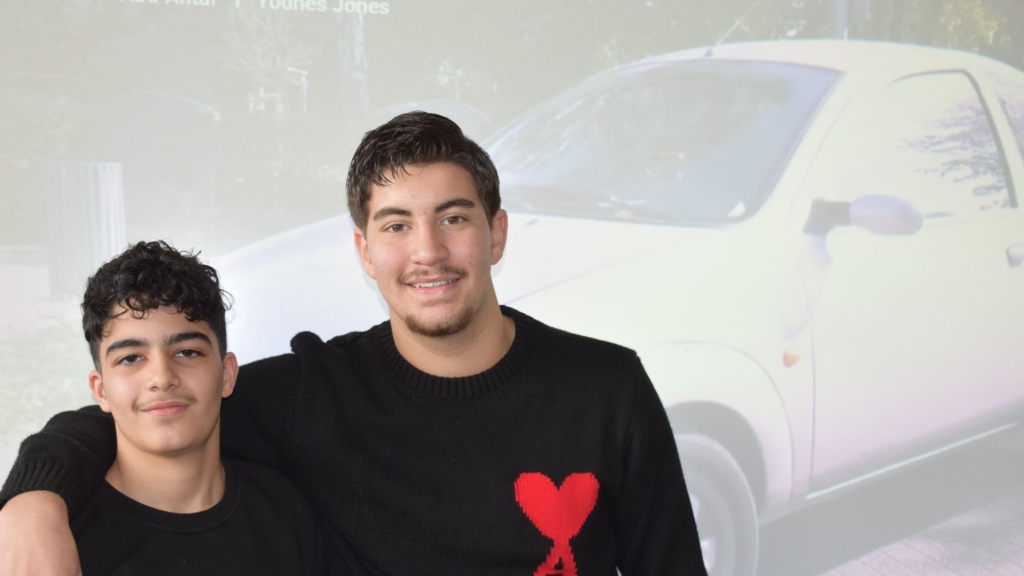 Zwei Jugendliche stehen eng nebeneinander vor einer Leinwand, die ein Auto zeigt.