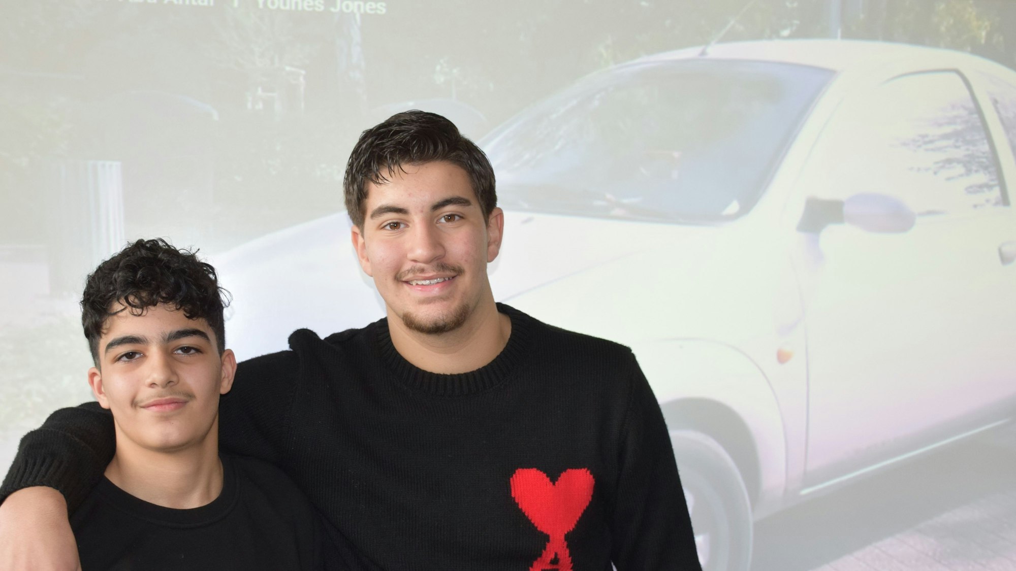 Zwei Jugendliche stehen eng nebeneinander vor einer Leinwand, die ein Auto zeigt.