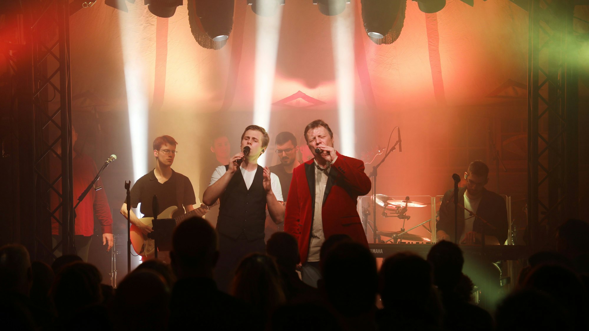 Zwei Sänger stehen im Scheinwerferlicht auf der Bühne gemeinsam mit einem Gitarristen, einem Keyborder und einem Schlagzeuger.