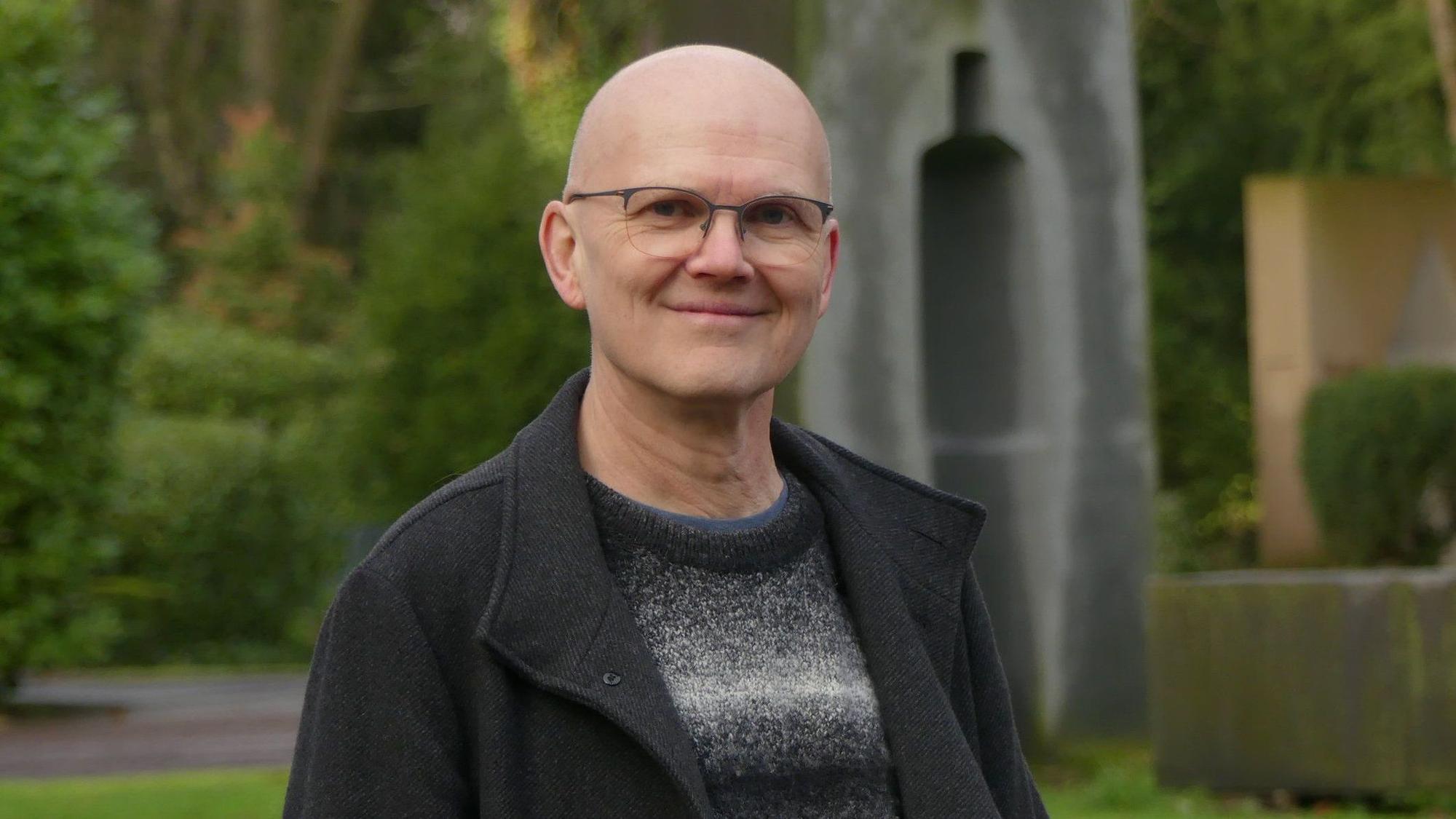 Ein Mann mit Glatze und Brille steht vor Grabmälern auf einem Friedhof.