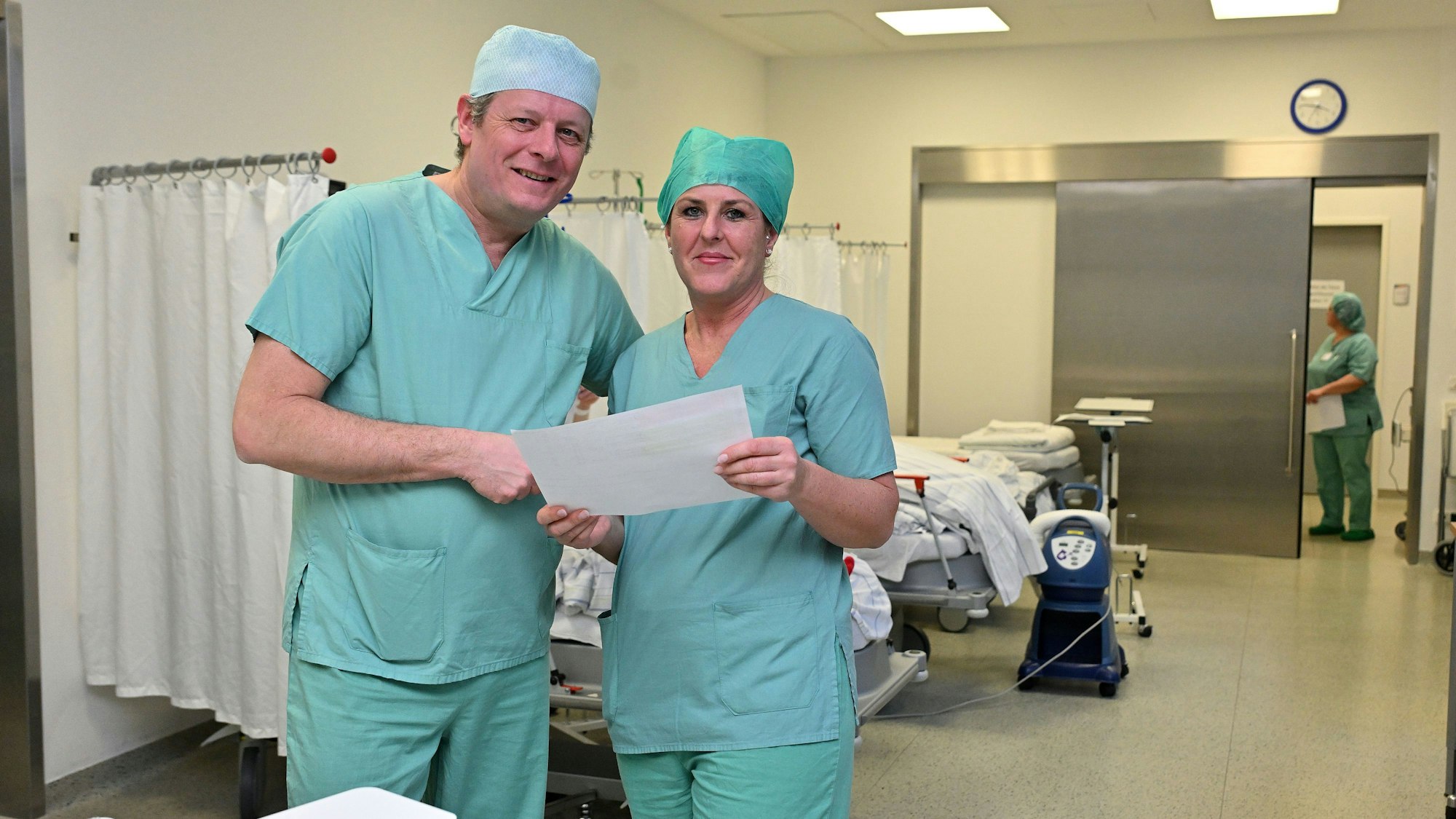 Chefarzt und OP-Managerin in OP-Kleidung stehen im Aufwachraum der Patienten und prüfen den Ausdruck für den heutigen OP-Plan.