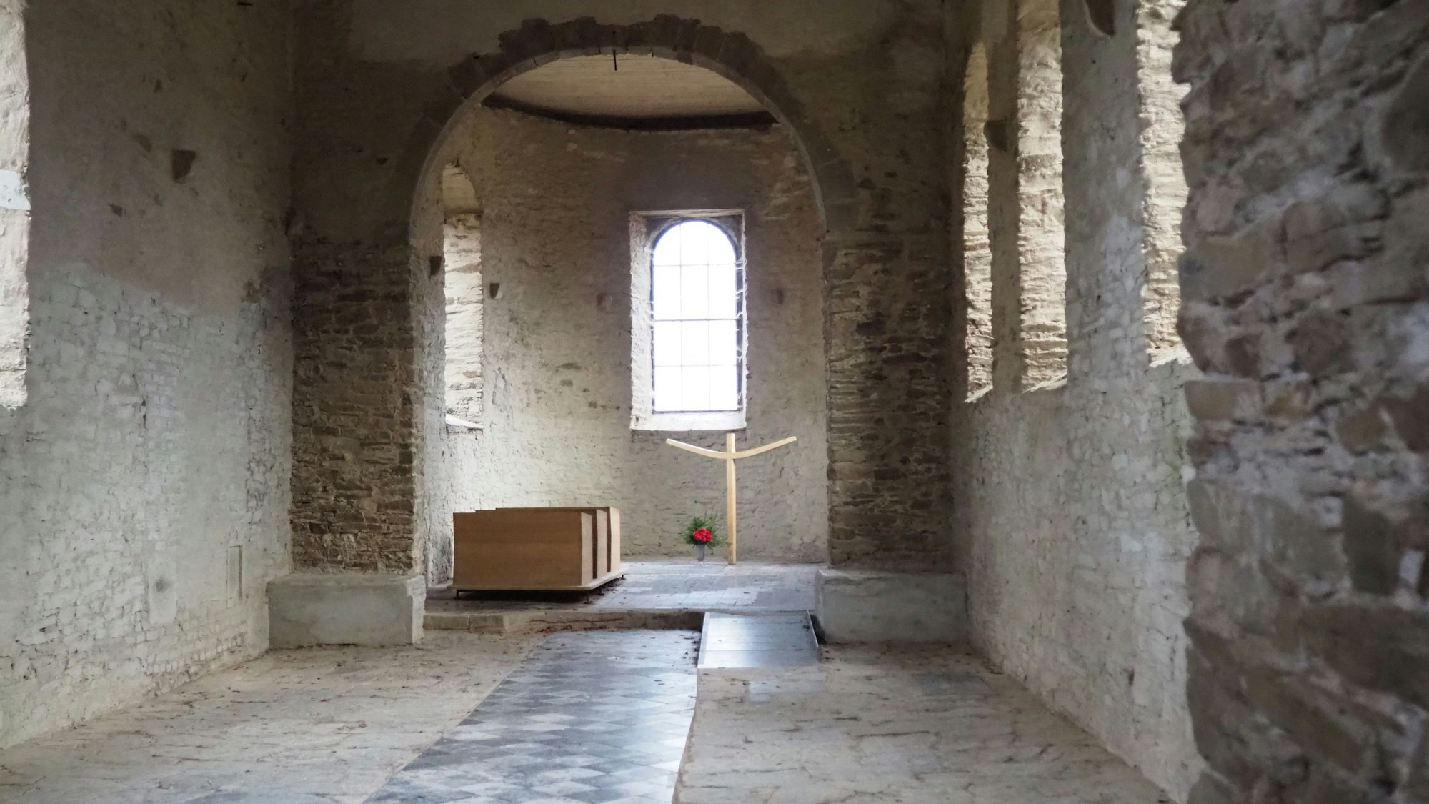 Blick in das unverputzte Kirchenschiff. Im Altarraum sind drei Sitzbänke und ein einfaches, modernes Holzkreuz zu sehen. Daneben eine Vase mit Blumen.