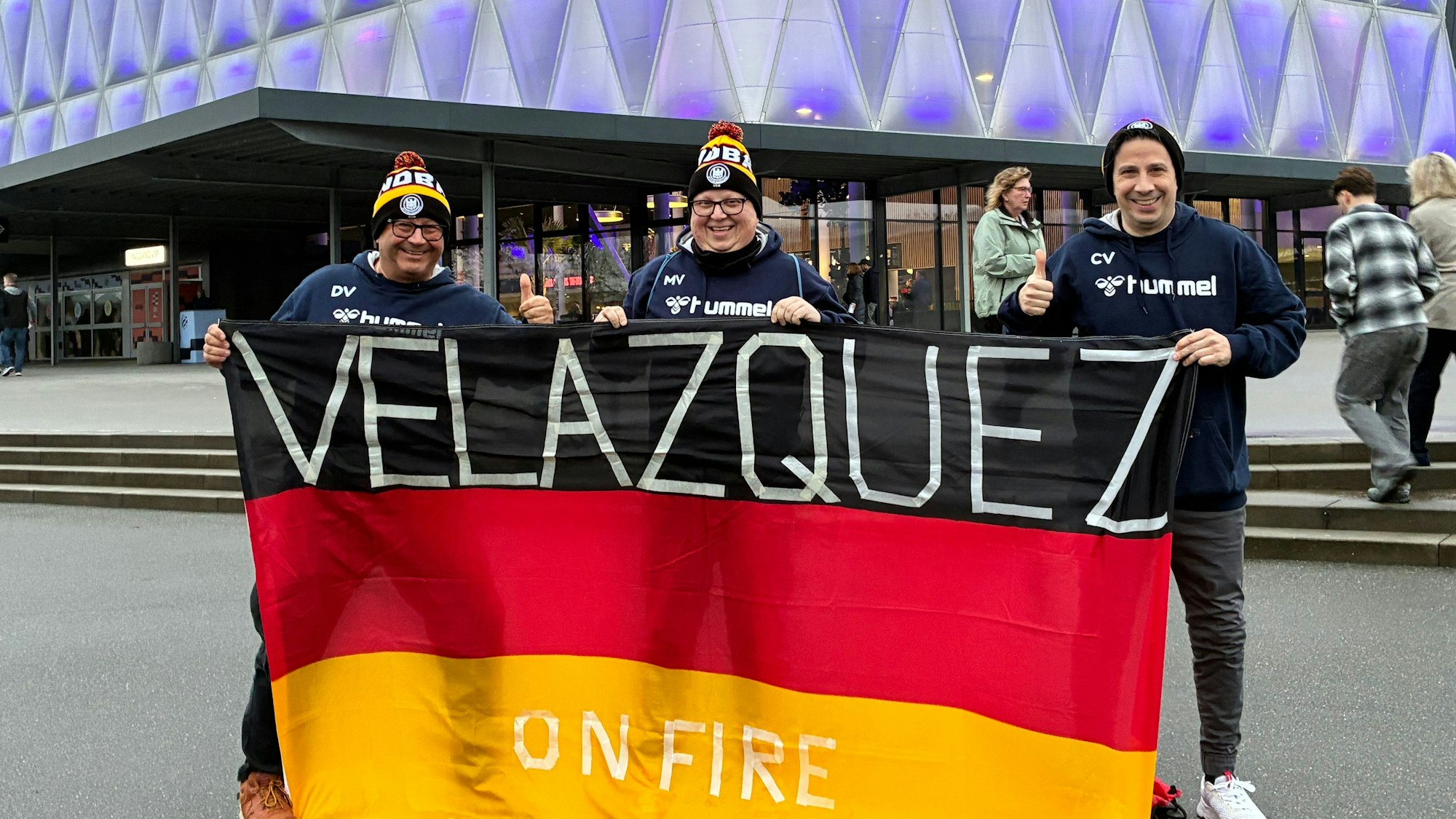 Drei Brüder stehen mit einer Deutschland-Fahne vor einer Handball-Halle.