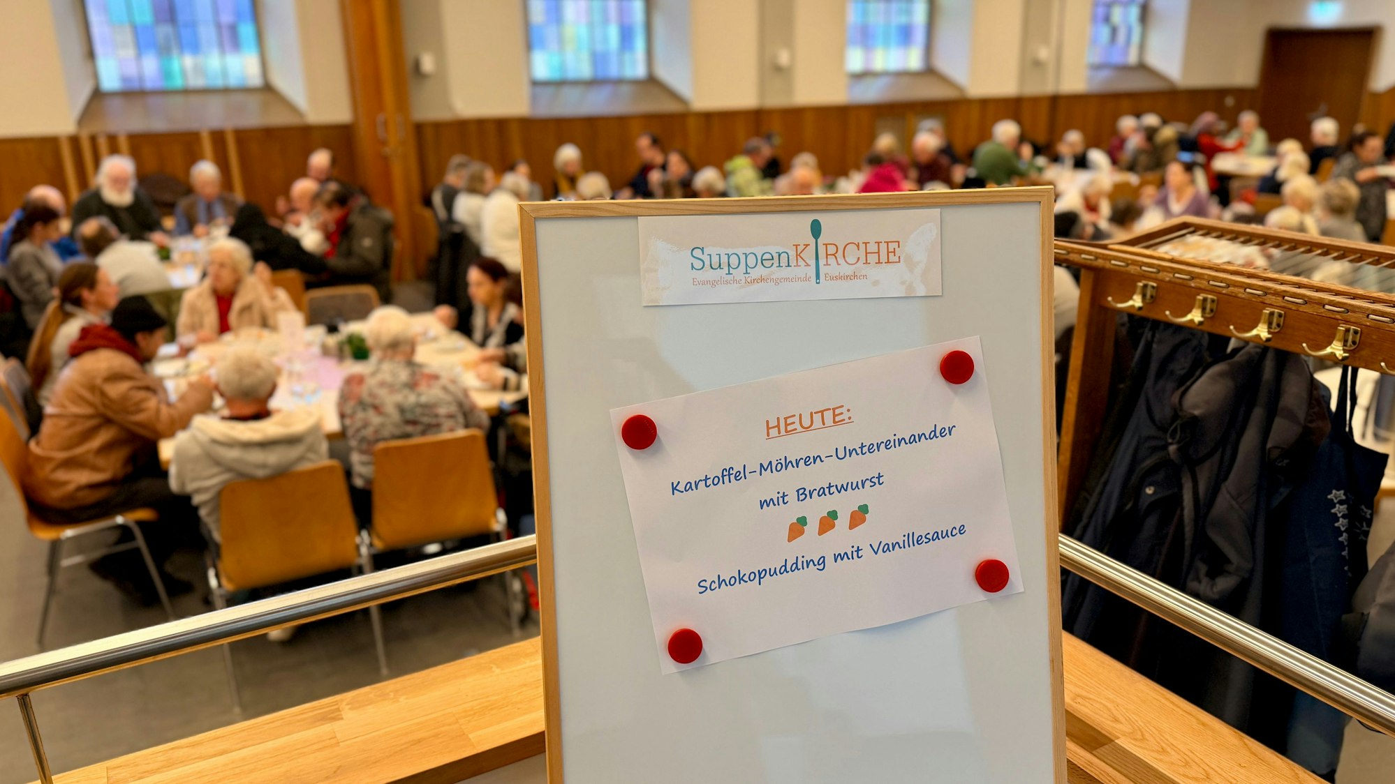 Am Eingang zum Gemeindesaal steht eine Tafel, auf der das Gericht des Tages angekündigt wird. Im Hintergrund sind die Tische bereits alle besetzt.