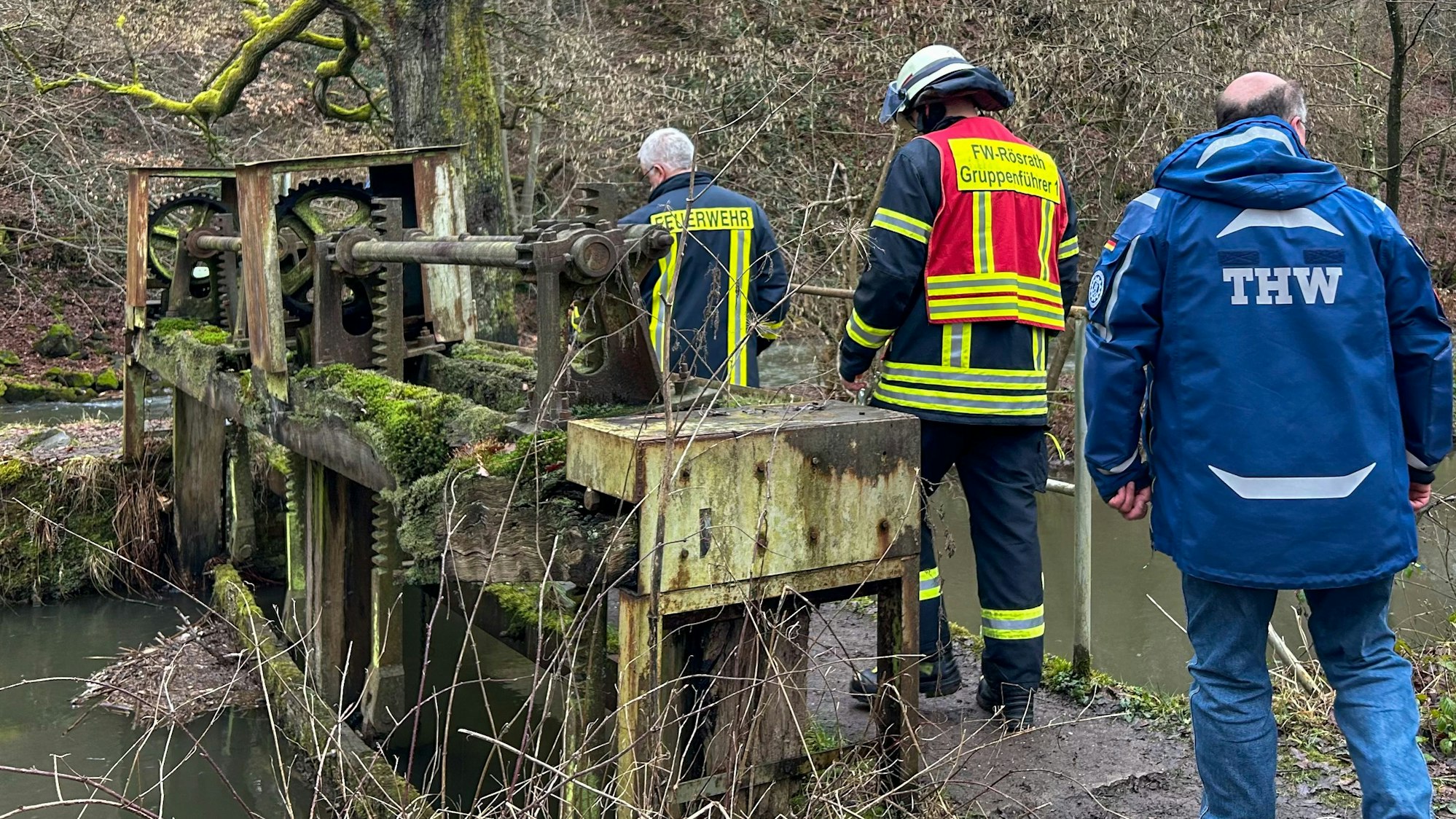 Feuerwehrleute und ein THW-Helfer gehen über ein marodes Absperrbauwerk am Wassergraben oberhalb des Sülzwehrs in Rösrath-Venauen.