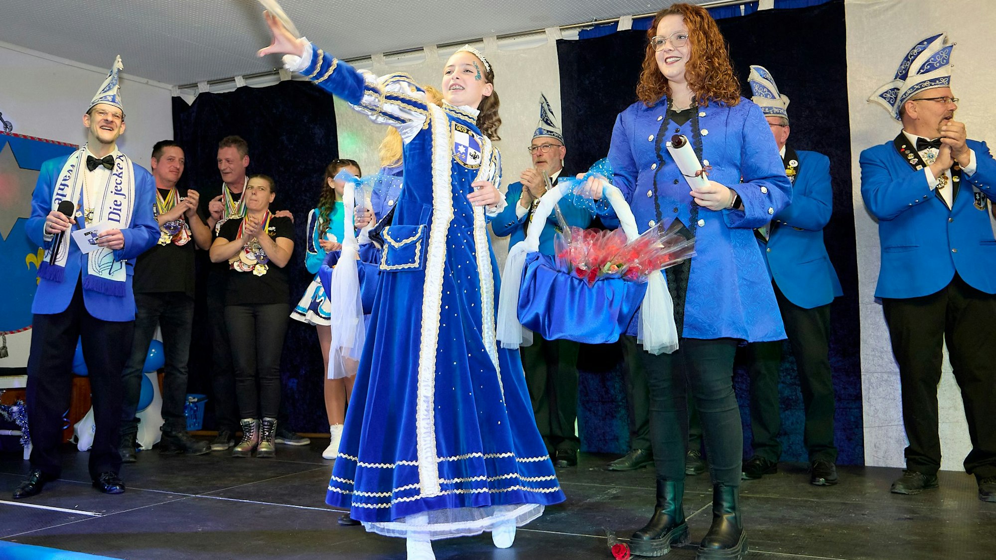 Prinzessin Mia hebt den rechten Arm zum Karnevalsgruß. Menschen auf der Bühne klatschen.