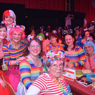 Ein Gruppe Frauen in gestreiften Oberteilen und angemalten Gesichtern sitzt an einem Biertisch in einem vollbesetzten Saal.
