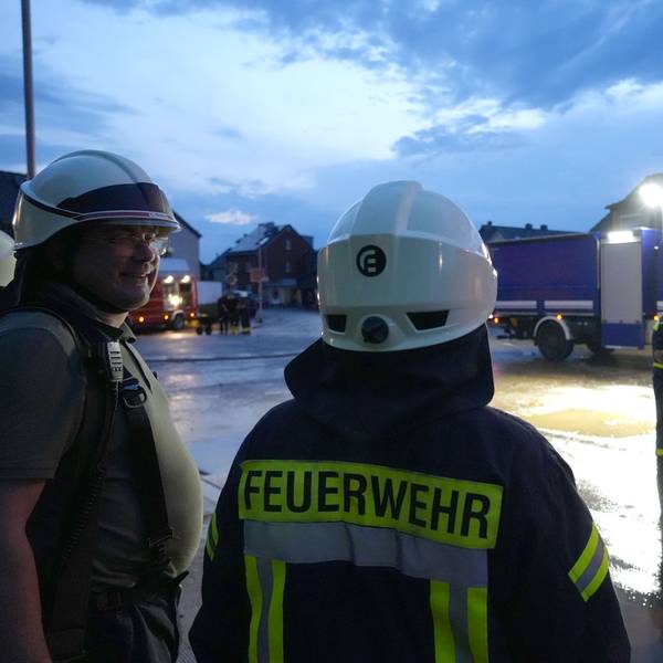 Zwei Feuerwehrmänner stehen an einer Straße, im Hintergrund sieht man weitere Feuerwehrleute sowie blaue Einsatzfahrzeuge.
