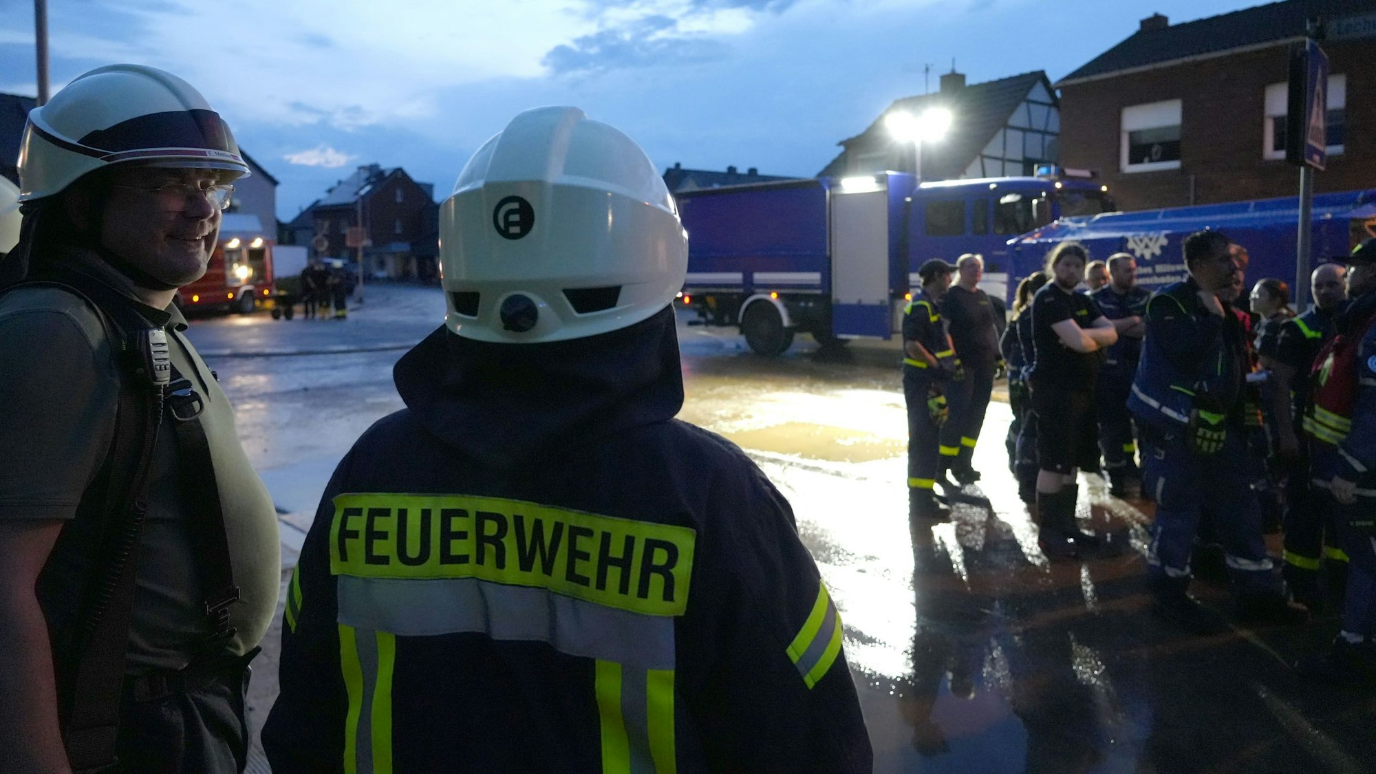 Zwei Feuerwehrmänner stehen an einer Straße, im Hintergrund sieht man weitere Feuerwehrleute sowie blaue Einsatzfahrzeuge.
