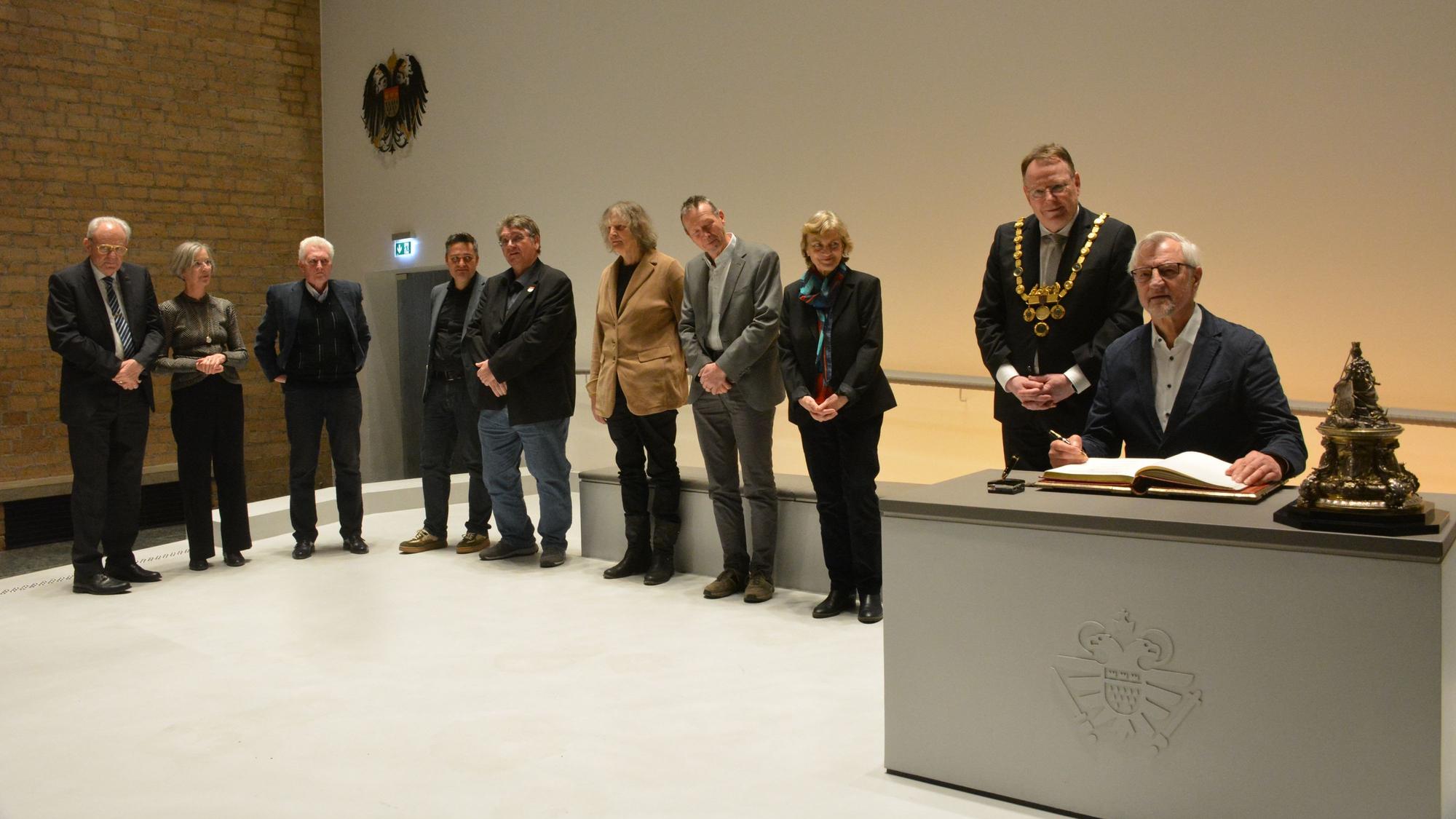 Ein Gruppe von acht Menschen steht in einer Reihe, ein Mann sitzt an einem Tisch, vor ihm liegt das Goldene Buch.