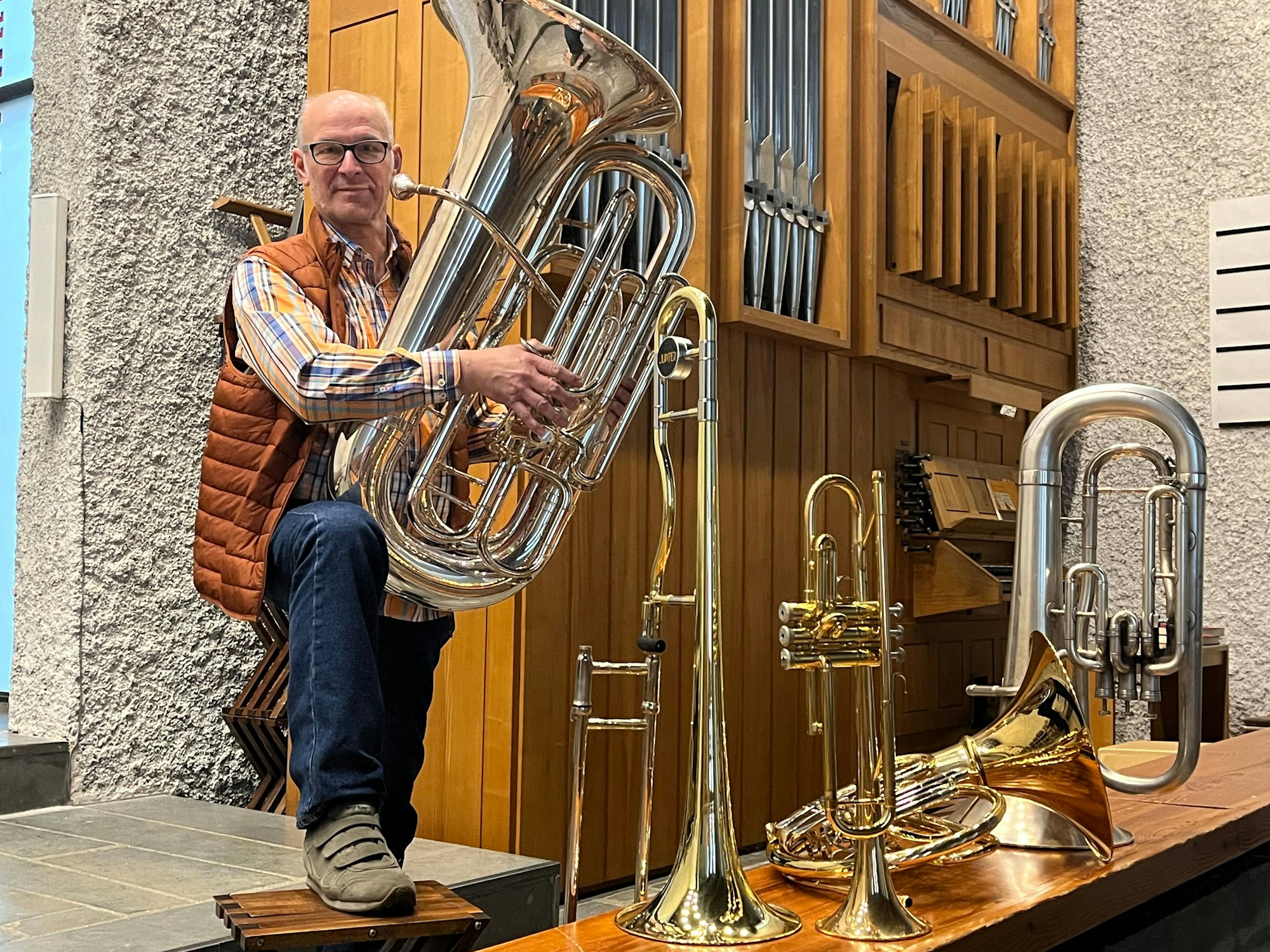 Ein Mann steht mit einer Tuba im Arm vor einer Orgel, vor ihm stehen weitere Blasinstrumente.