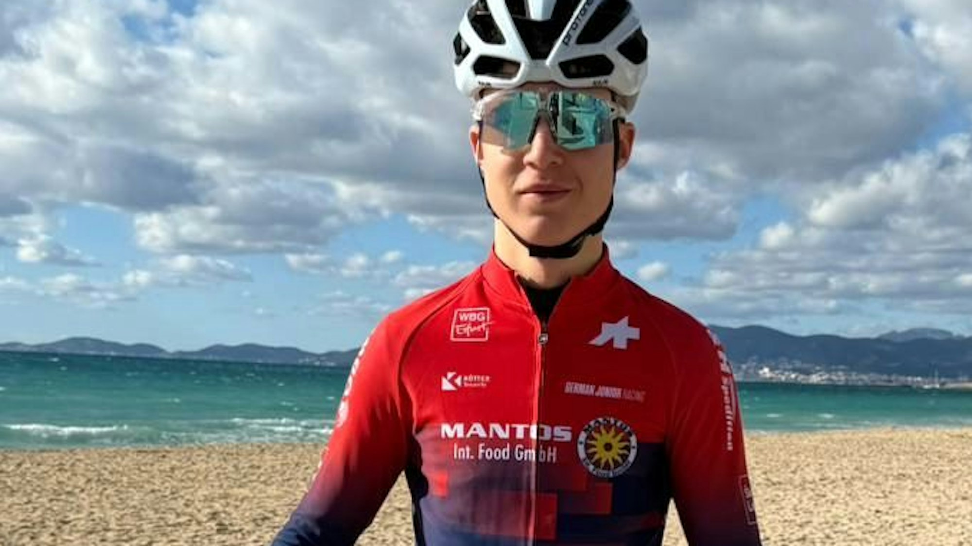 Mika Lohmar steht am Strand von Mallorca und hält sein Fahrrad in den Händen.