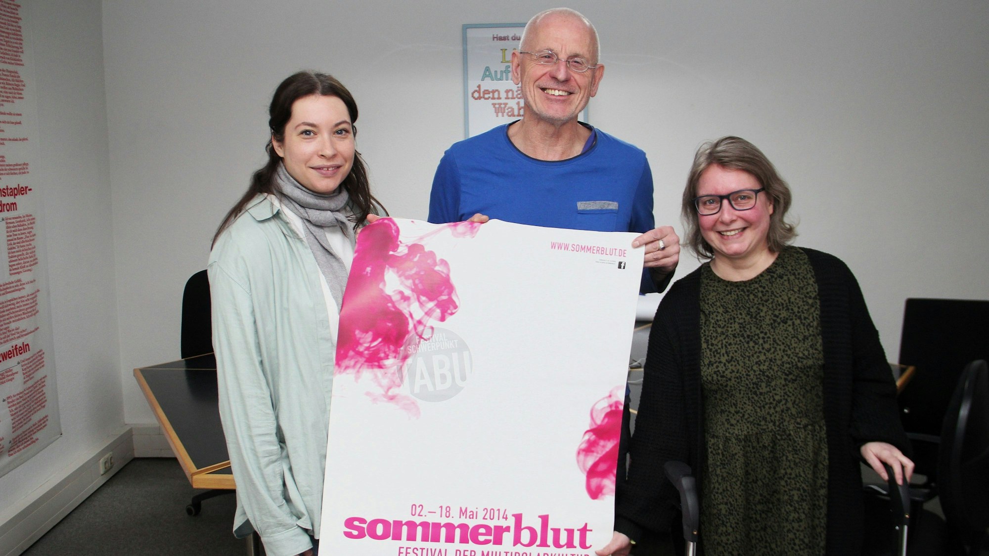 Zwei Frauen und ein Mann stehen nebeneinander. Der Mann hält ein Plakat, auf dem „Sommerblut“ zu lesen ist.