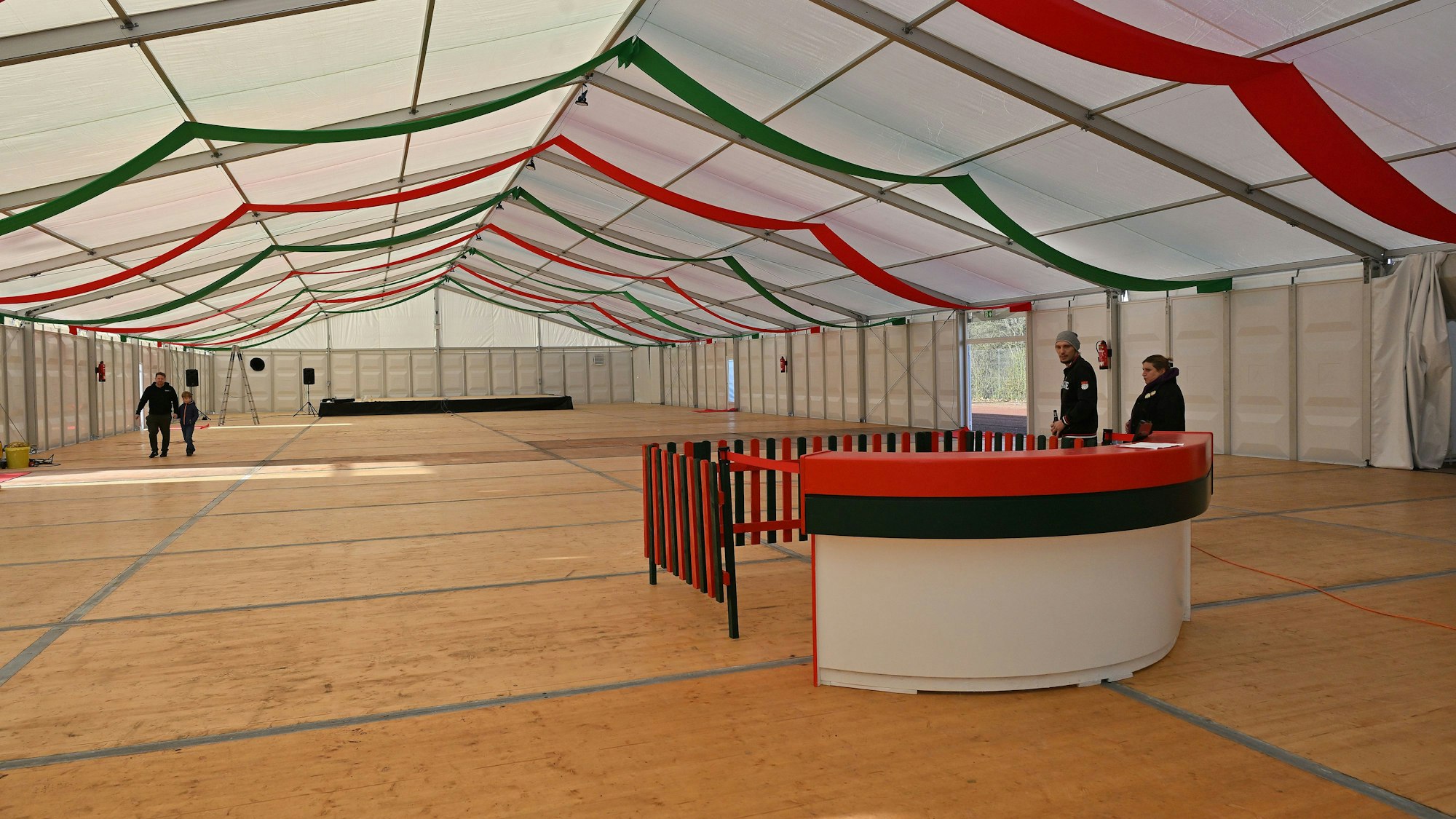 Ein leeres Festzelt mit Holzboden und einer Theke in Grün-Rot, unter dem weißen Zeltdach sind rot-grüne Stoffbahnen gespannt.