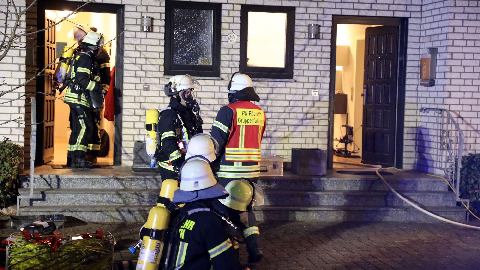 Feuerwehrleute kommen aus der Tür eines Wohnhauses, andere stehen davor auf einem gepflasterten Platz.