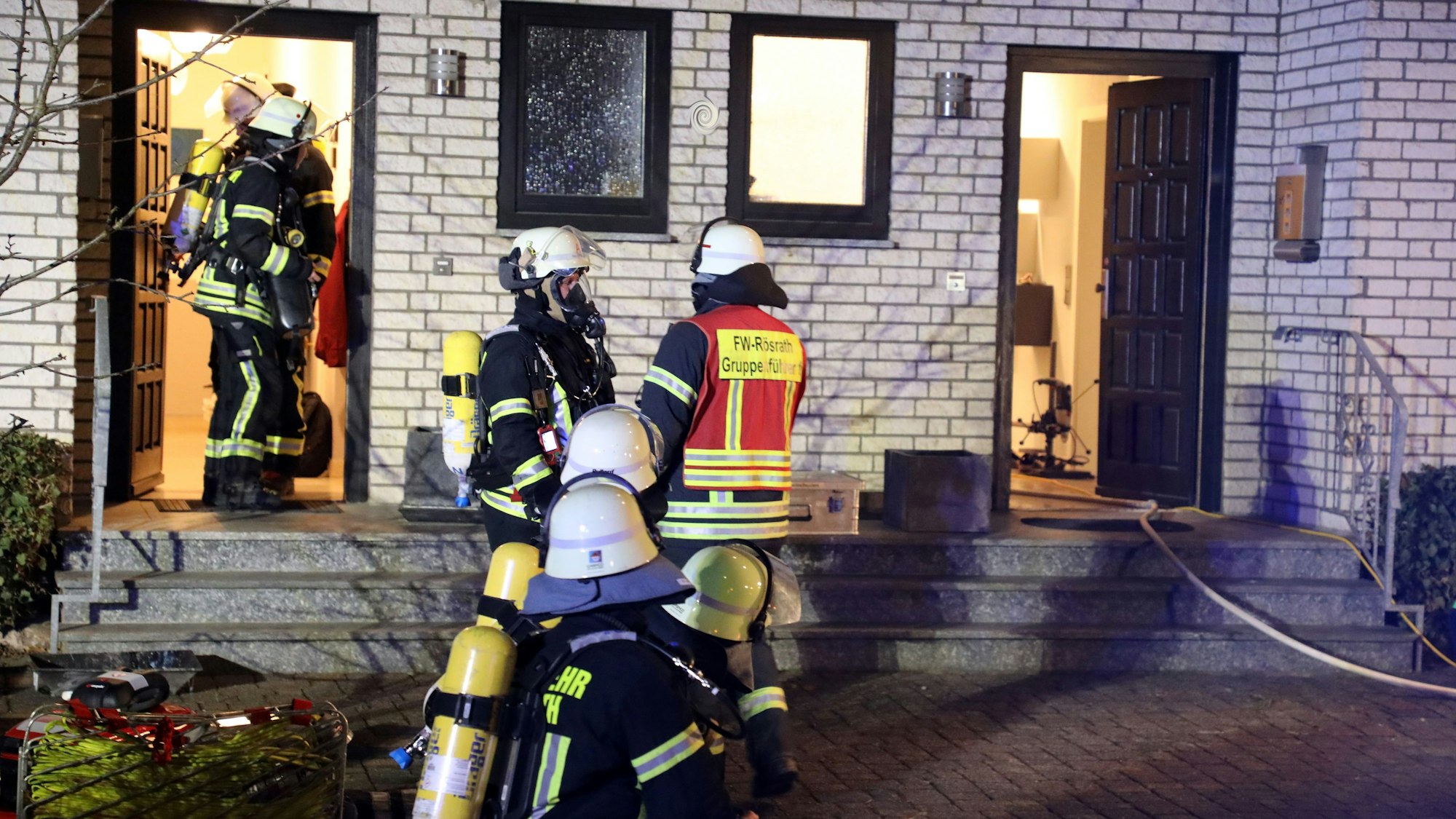 Feuerwehrleute kommen aus der Tür eines Wohnhauses, andere stehen davor auf einem gepflasterten Platz.