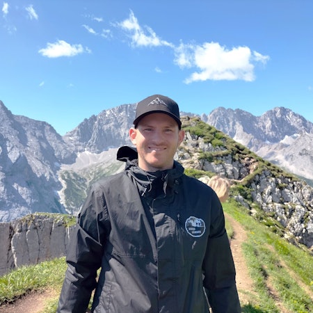 Ein junger Mann mit Kappe auf dem Kopf steht vor einem Bergpanorama.