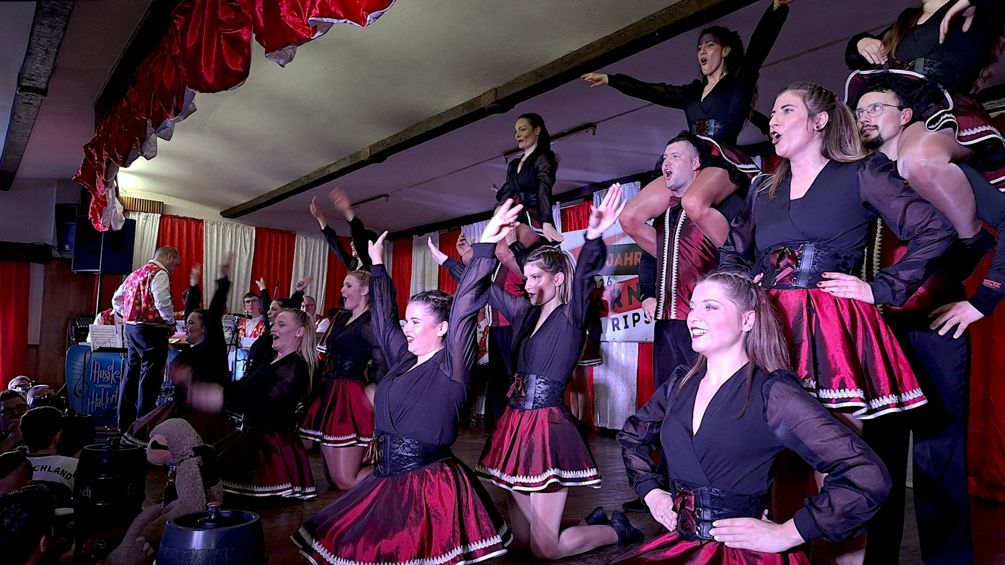 In rot-schwarzen Kleidern stehen die Tänzer und Tänzerinnen auf der Bühne. Manche machen Hebefiguren.