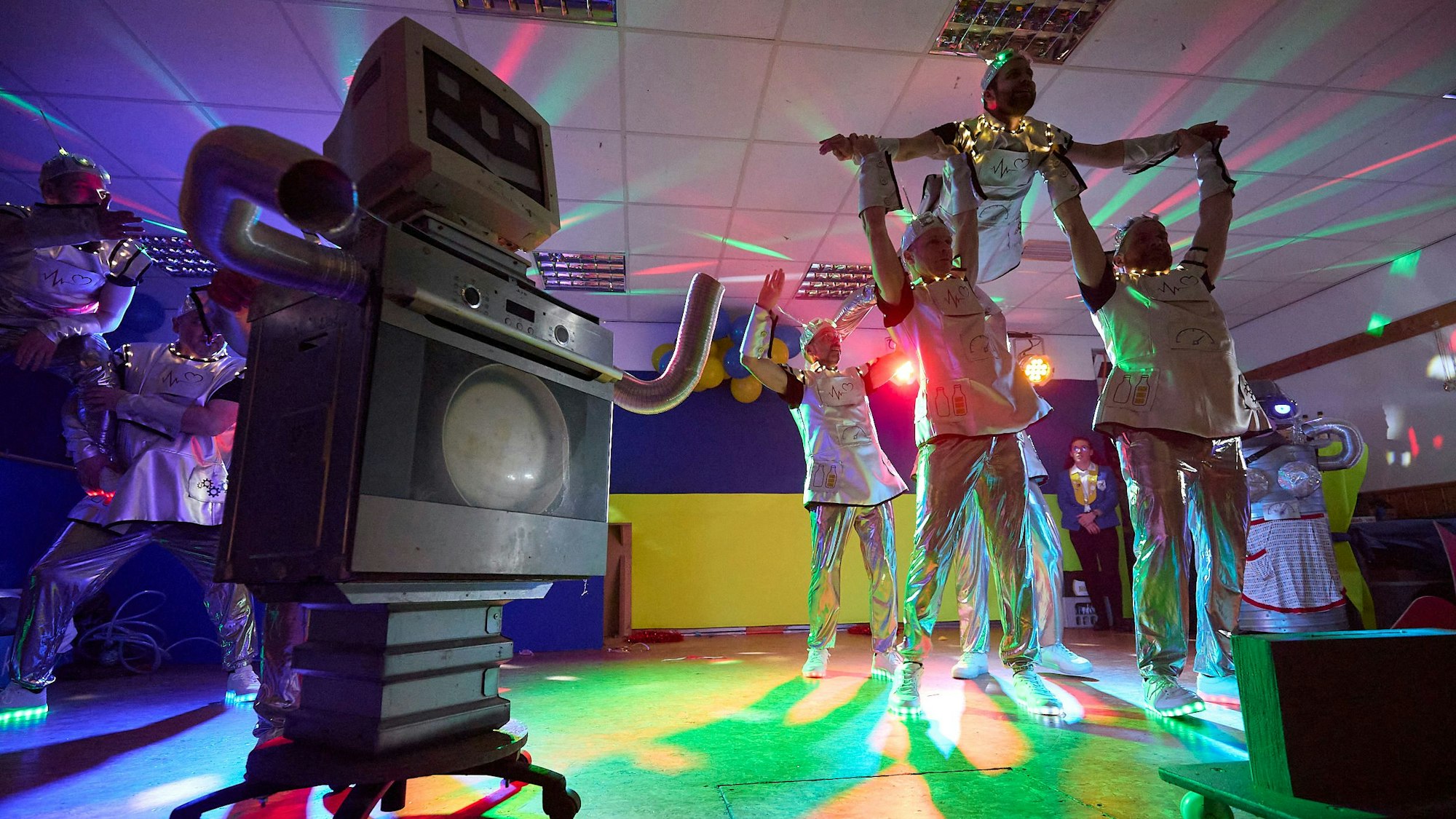 In einem Raum mit Discolicht machen kostümierte Männer eine Hebefigur. Im Vordergrund des Bildes steht ein Küchenherd, der zum Roboter umgebaut wurde.