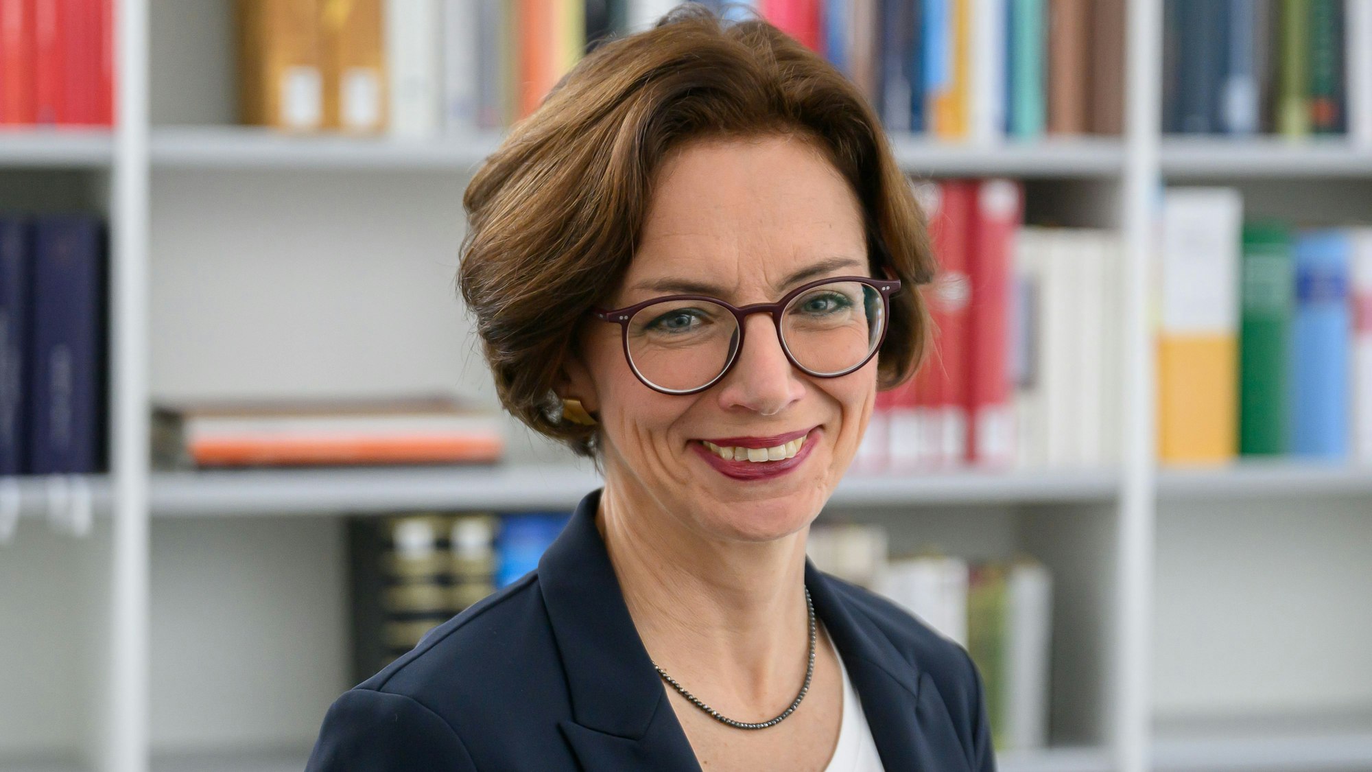 Eine Frau mit braunen Haaren und Brille vor einem Bücherregal.