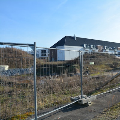 Zu sehen ist ein Bauzaun vor einer Baugrube, dahinter ein Gebäudekomplex.