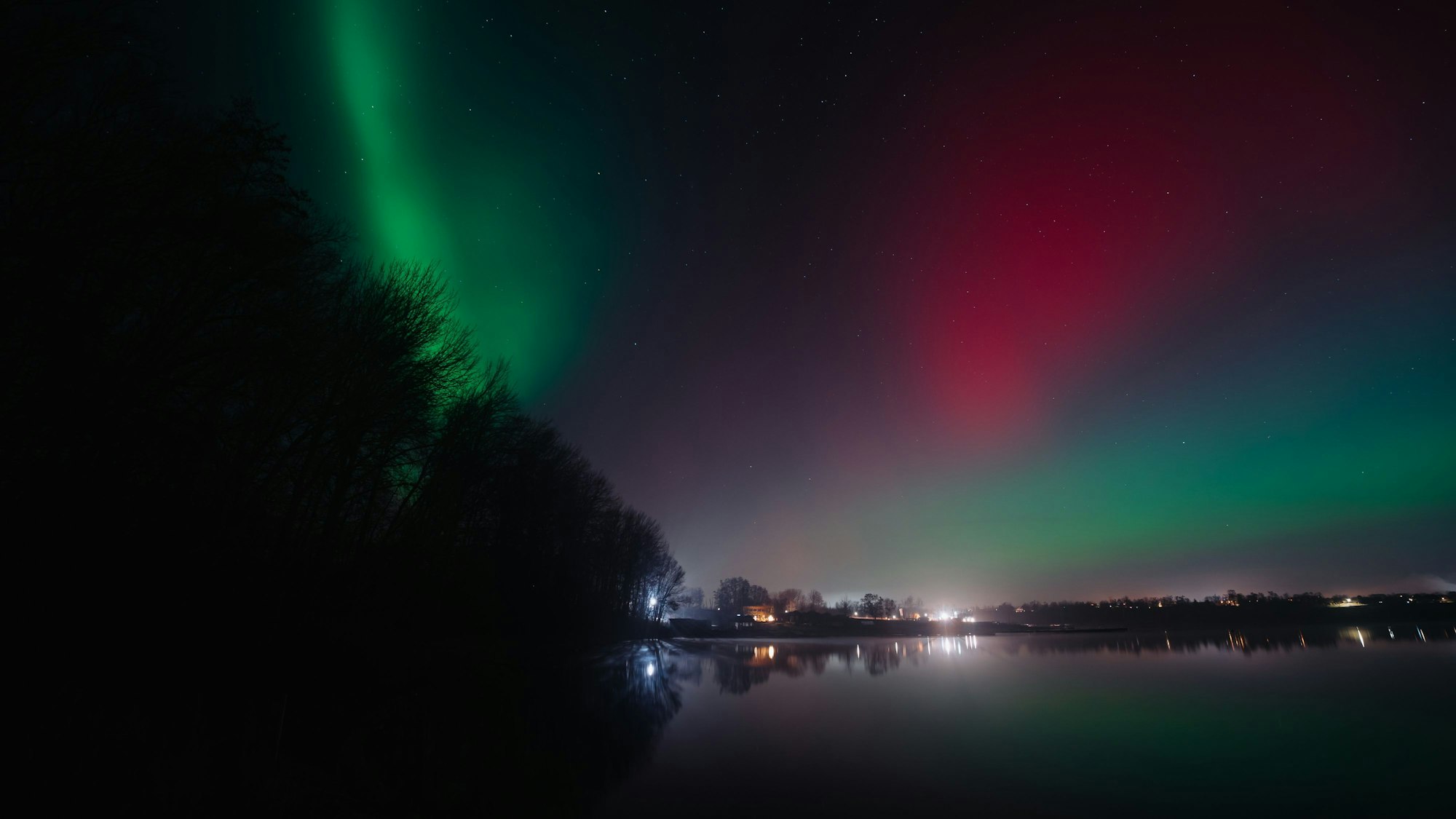 Über den Zülpicher See bringen Polarlichter ein bisschen Helligkeit in den dunklen Nachthimmel. Auf der Seeoberfläche spiegeln sich Gebäude und Bäume.