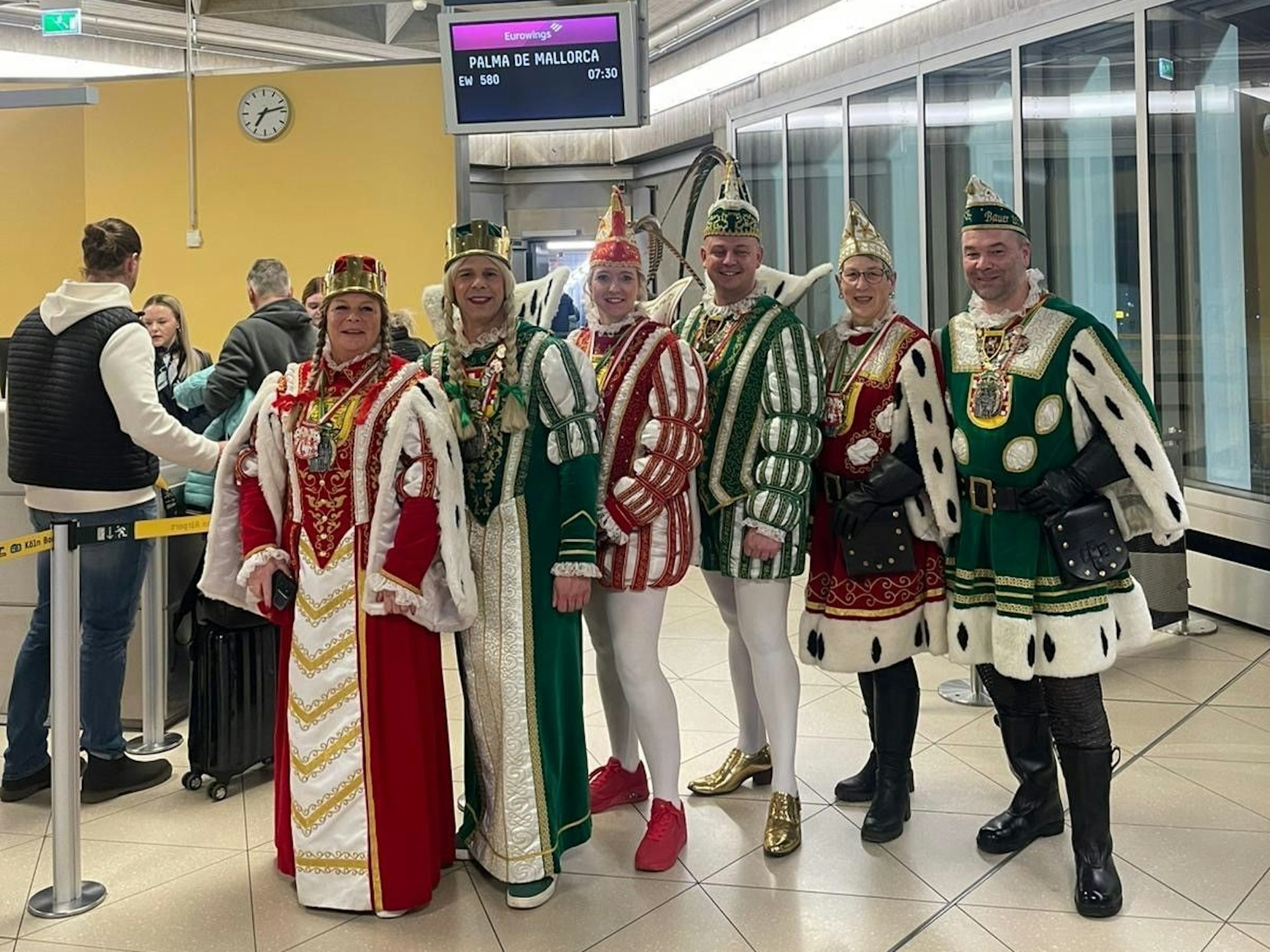 Karnevalisten stehen an einem Flughafengate. Die Anzeige im Hintergrund zeigt als Ziel Palma de Mallorca.
