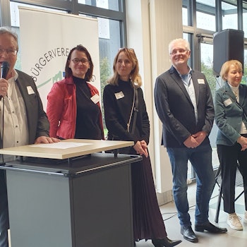 Sieben Personen stehen nebeneinander. Der Mann ganz links steht an einem Rednerpult und hält ein Mikrofon.