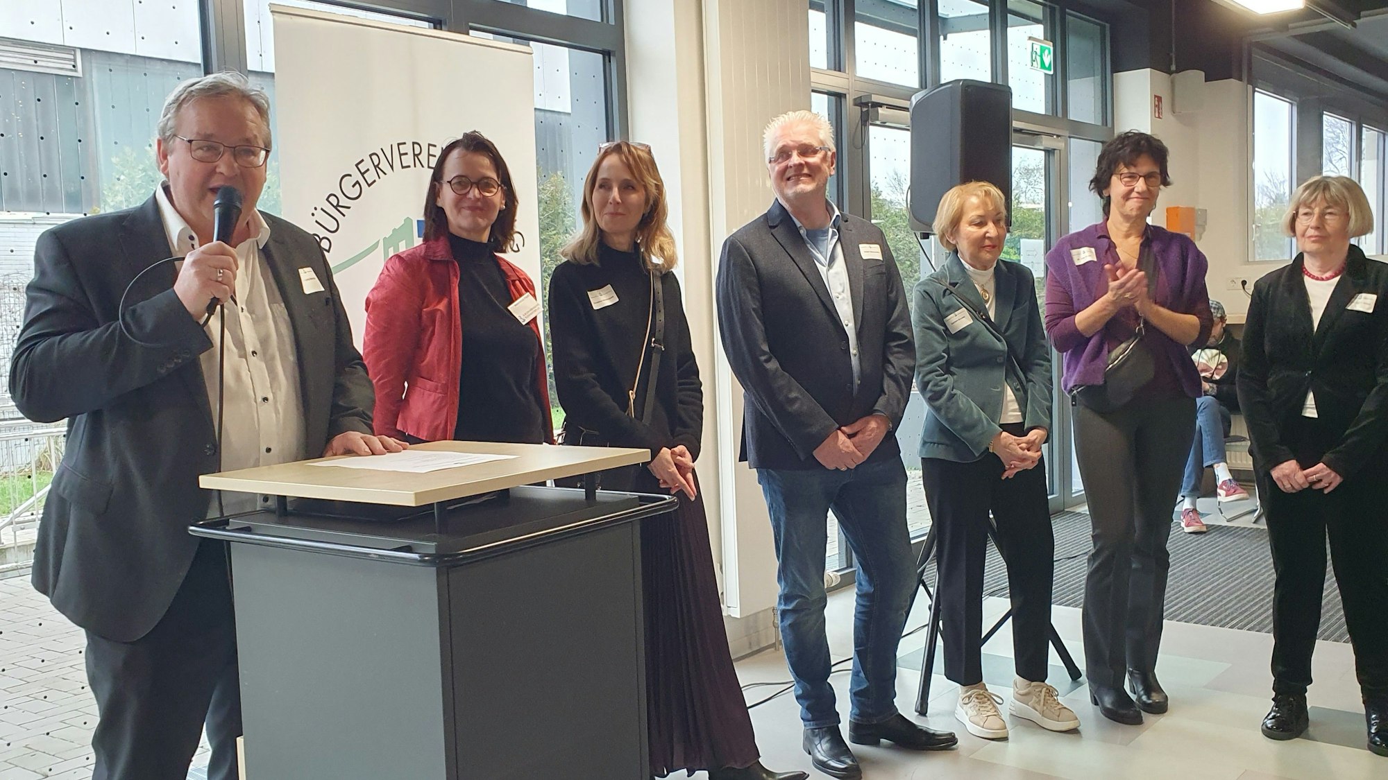 Sieben Personen stehen nebeneinander. Der Mann ganz links steht an einem Rednerpult und hält ein Mikrofon.