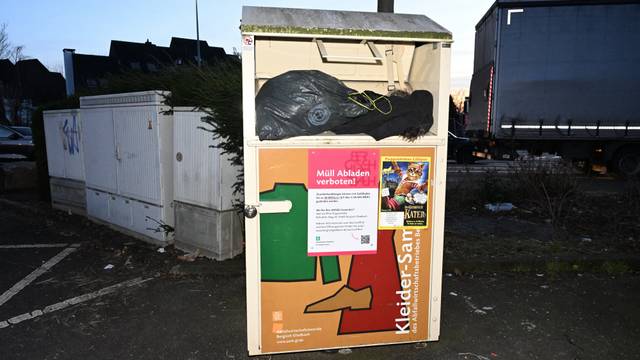 Ein Altkleidercontainer, aus dem volle schwarze Plastikäcke hervorquellen, vor einem Stromkasten.