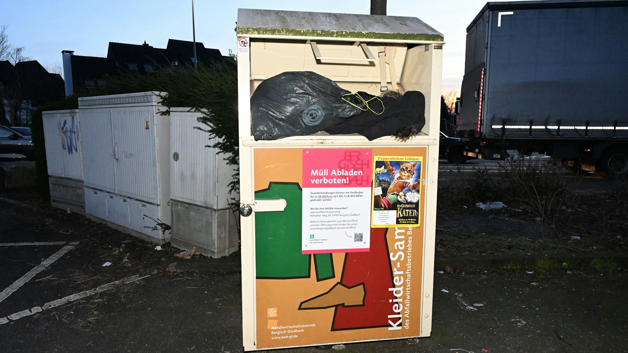 Ein Altkleidercontainer, aus dem volle schwarze Plastikäcke hervorquellen, vor einem Stromkasten.