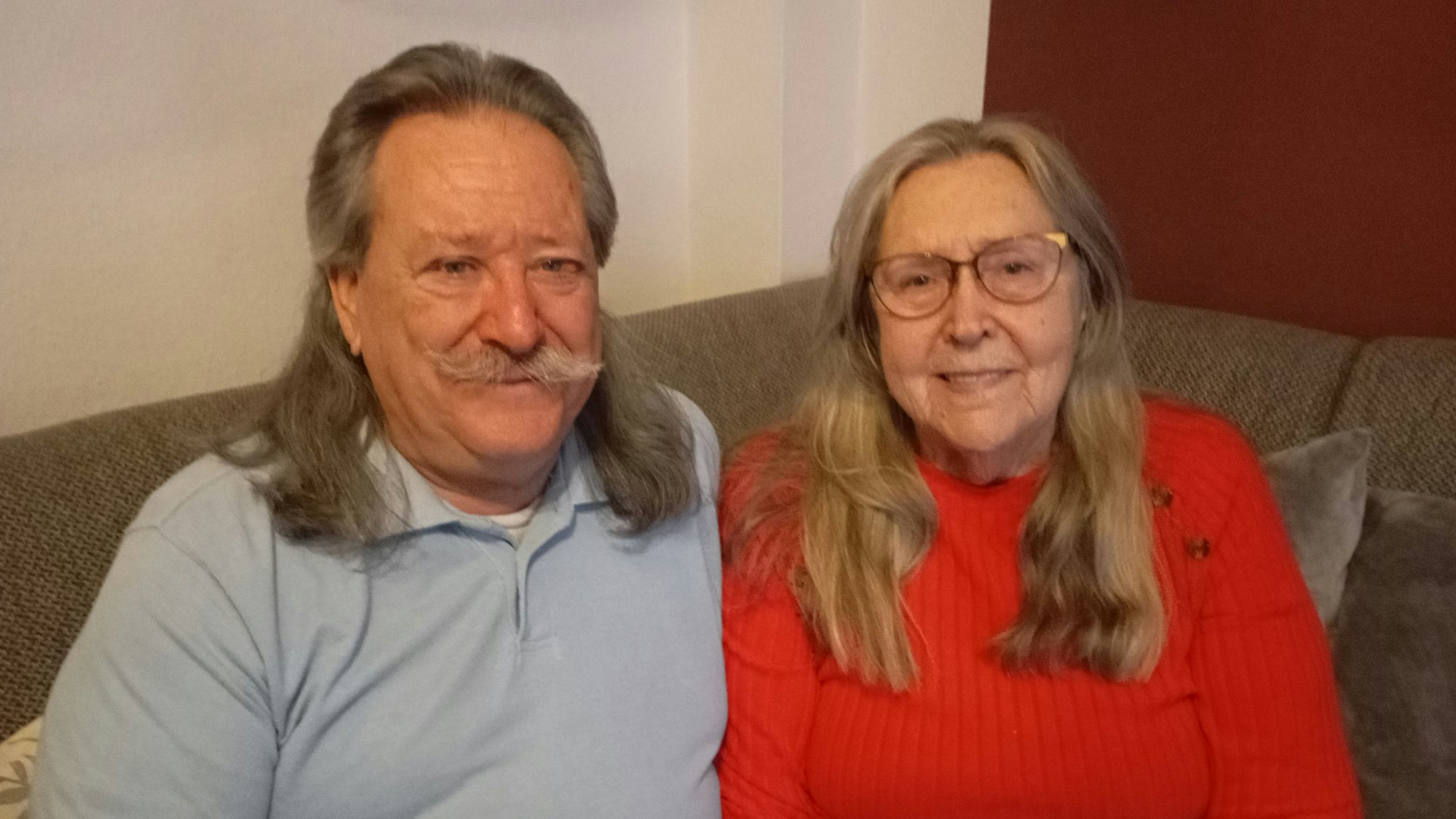 Mario Capua trägt ein blaues Polo-Hemd. Er hat lange Haare und trägt einen Schnurbart. Elisabeth Capua trägt eine Brille und einen roten Pullover.