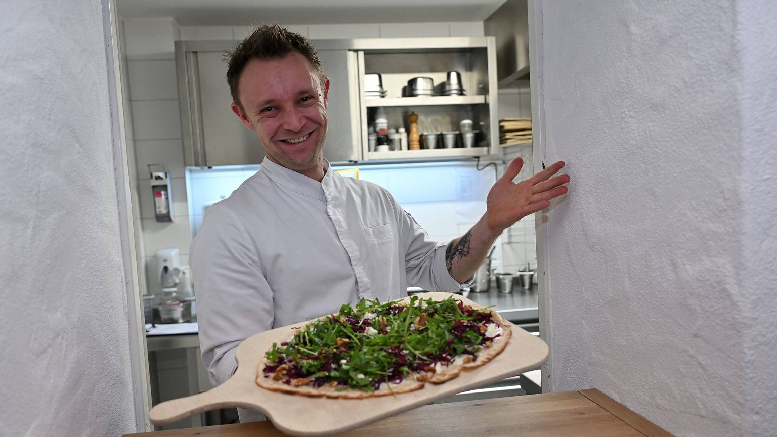Ein lachender Koch in weißer Jacke reicht dem Betrachter einen Flammkuchen mit grünem Salat auf einem großen Holzbrett entgegen.