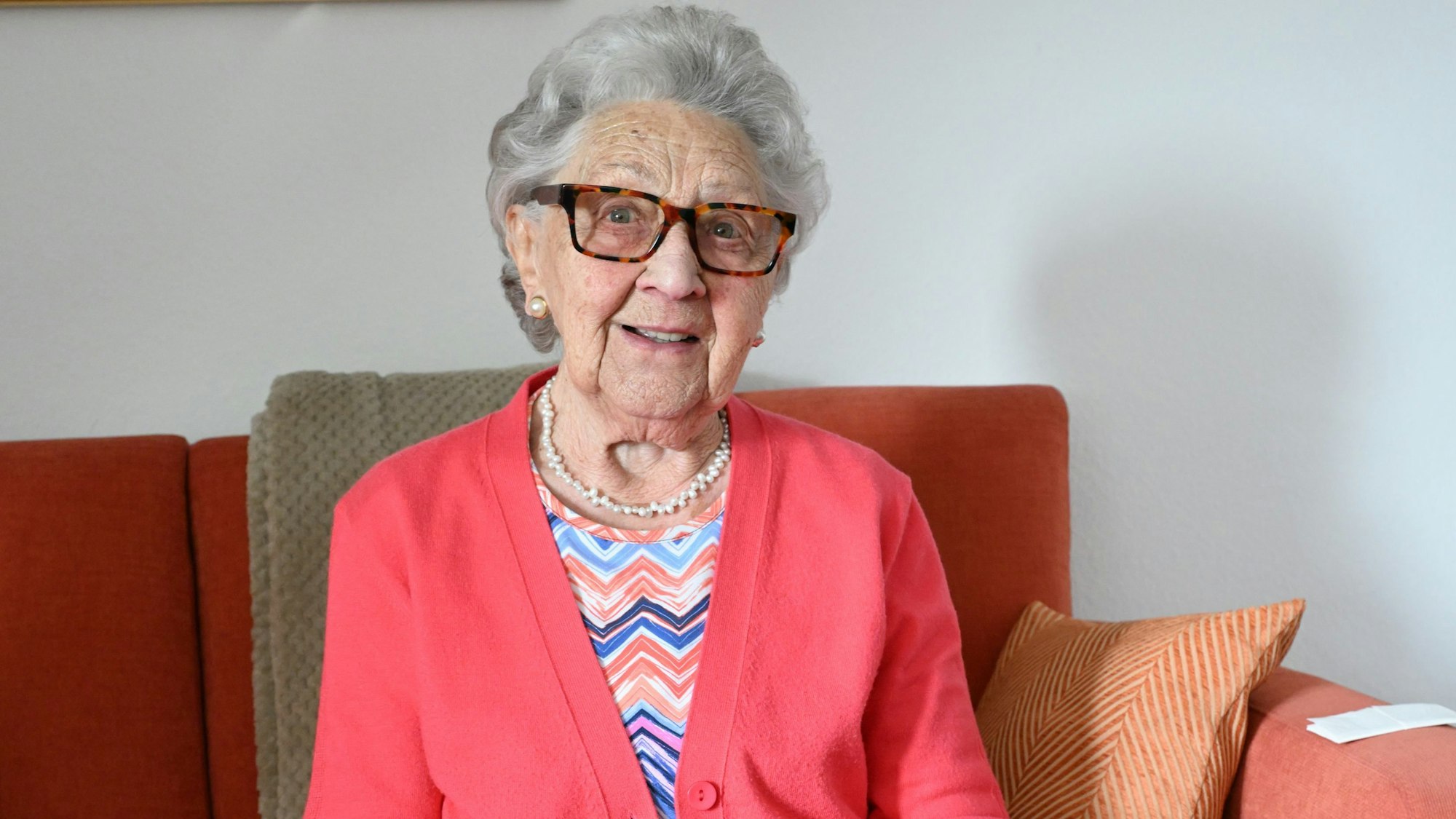 Bündgen trägt graue gelockte Haare, eine Brille und einen pinken Blazer, während sie auf der Couch sitzt.