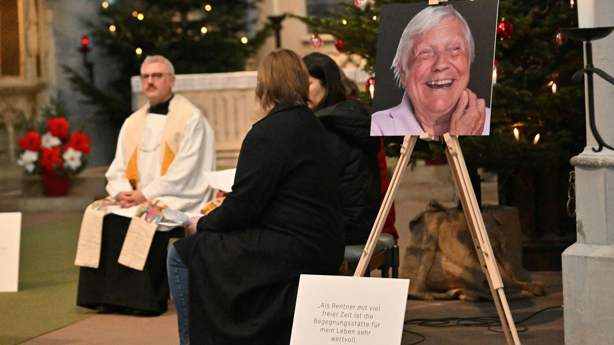 Ein katholischer Pfarrer sitzt im Altarraum einer Kirche, seitlich ist ein großes Foto aufgestellt, es zeigt das Gesicht eines lachenden Mannes.