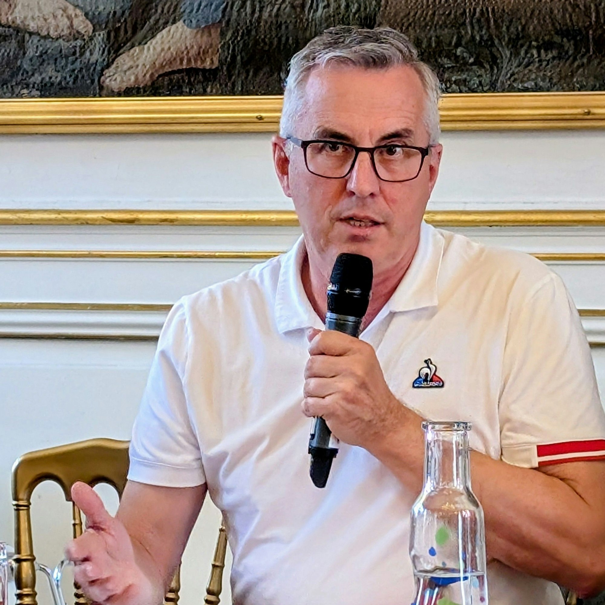 Andreas Wolter trägt ein weißes Poloshirt und spricht in ein Mikrofon, das er in der rechten Hand hält.