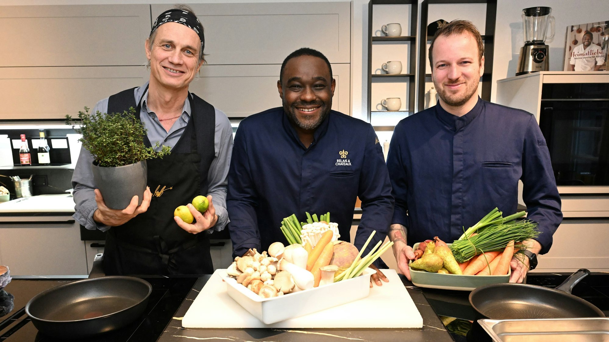 Schauspieler Ralf Bauer und den Köchen Nelson Müller und Jordy van der Ven stehen in einer Küche, vor ihnen liegt Gemüse wie Mähren und Inger sowie verschiedene Pilzsorten (v.l.n.r.).