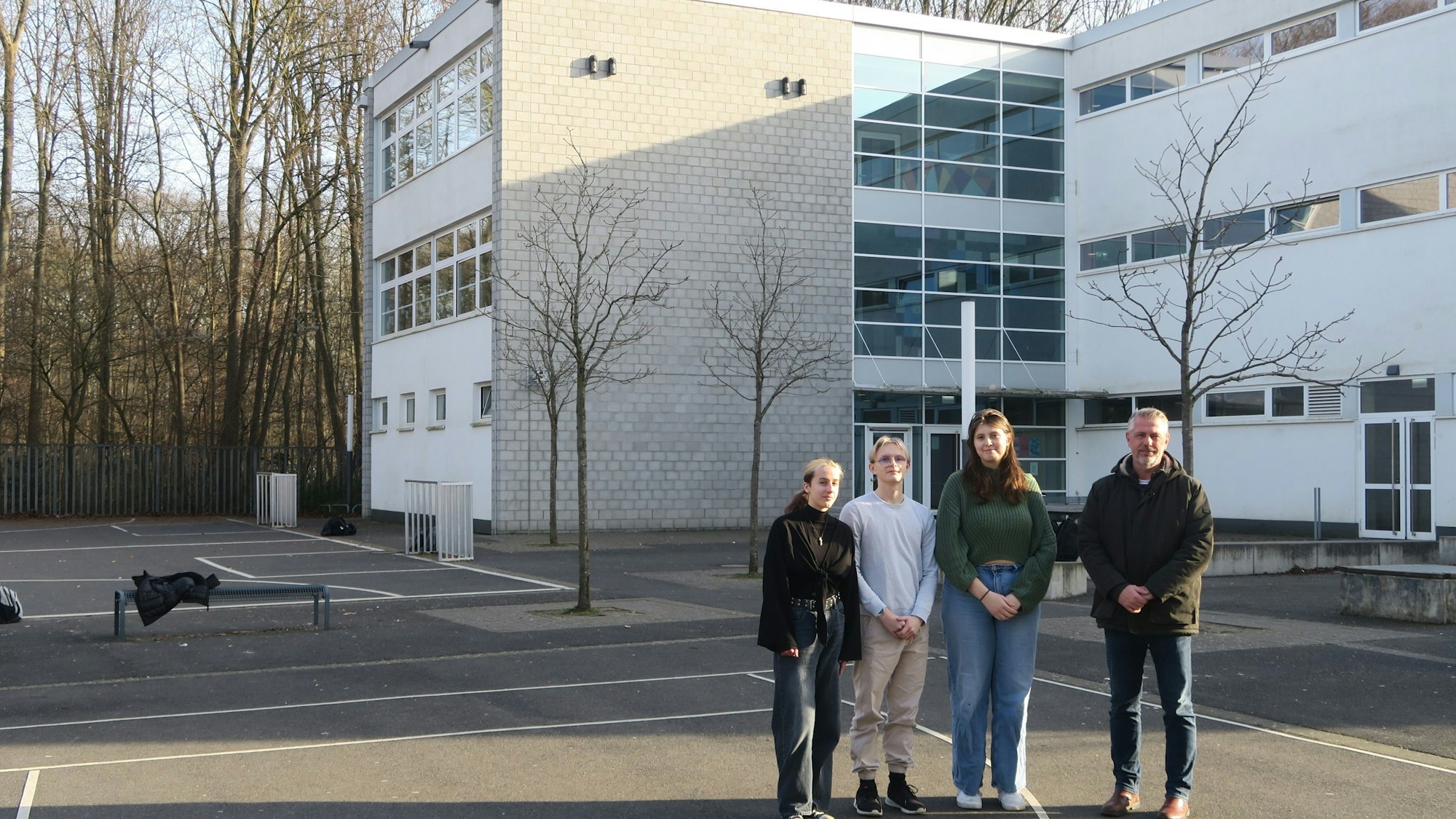 Drei Schüler und ein Lehrer stehen auf einem großen asphaltierten Schulhof.