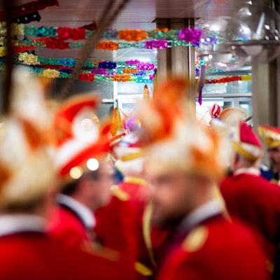 In einem mit Girlanden dekorierten Saal sind Karnevalisten mit roten Jacketts und Narrenkappen unscharf zu sehen.