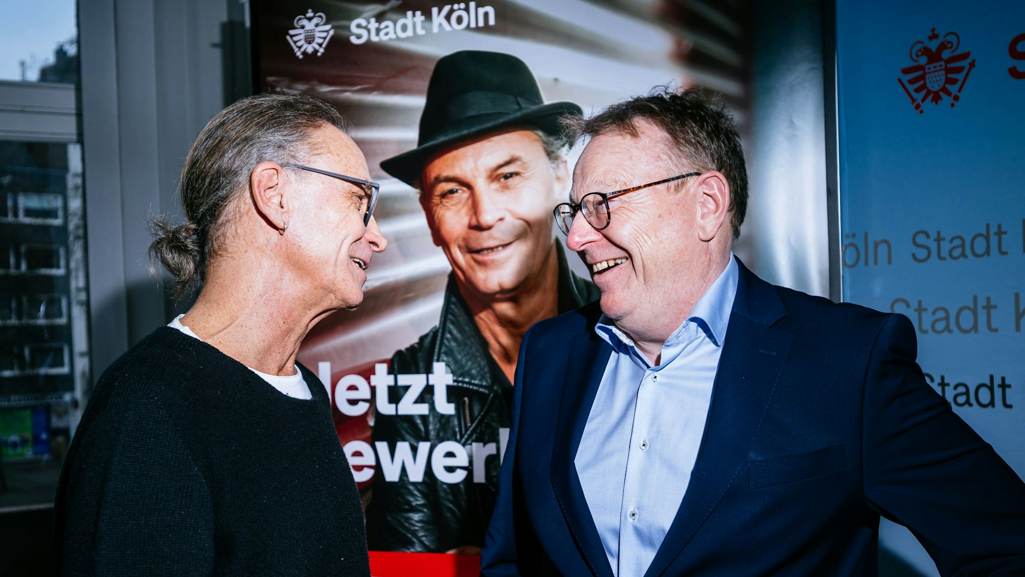 Der Ehrenamtspate Peter Brings steht links, rechts steht Oberbürgermeister Torsten Burmester. Die beiden lachen sich gegenseitig an, die Gesichter sind vom Profil aus zu sehen. Sie stehen vor dem Werbeschild zu dem Ehrenamtspreis.