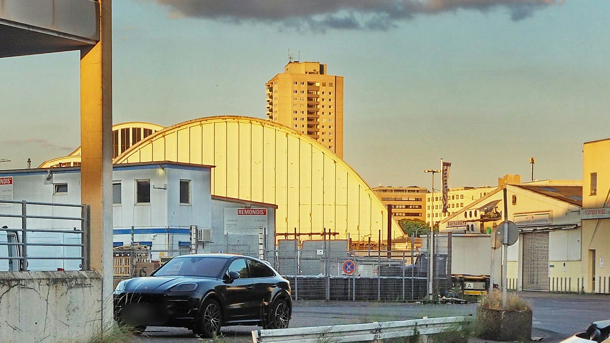 Vor einer großen Halle stehen verschiedene Container-Bauten, es sind Bauzäune und ein dunkler SUV zu sehen.