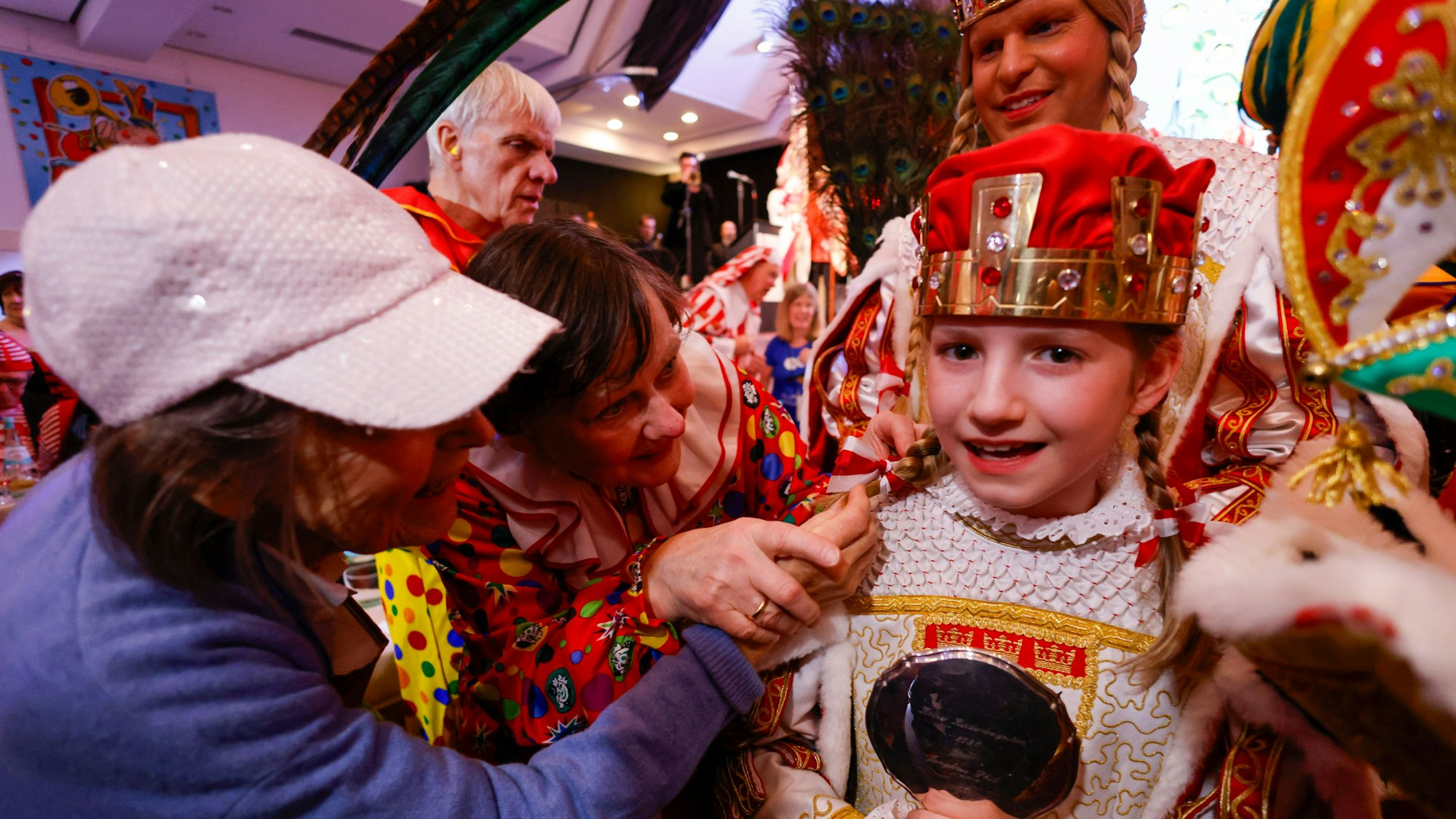 Die Jungfrau des Kinderdreigestirns Philomena Jürgens. Zwei Personen ertasten einen ihrer zwei blonden Zöpfe ab. Hinter Philomena steht Stefan Blatt, Jungfrau des großen Dreigestirns. Philomena lächelt in die Kamera.