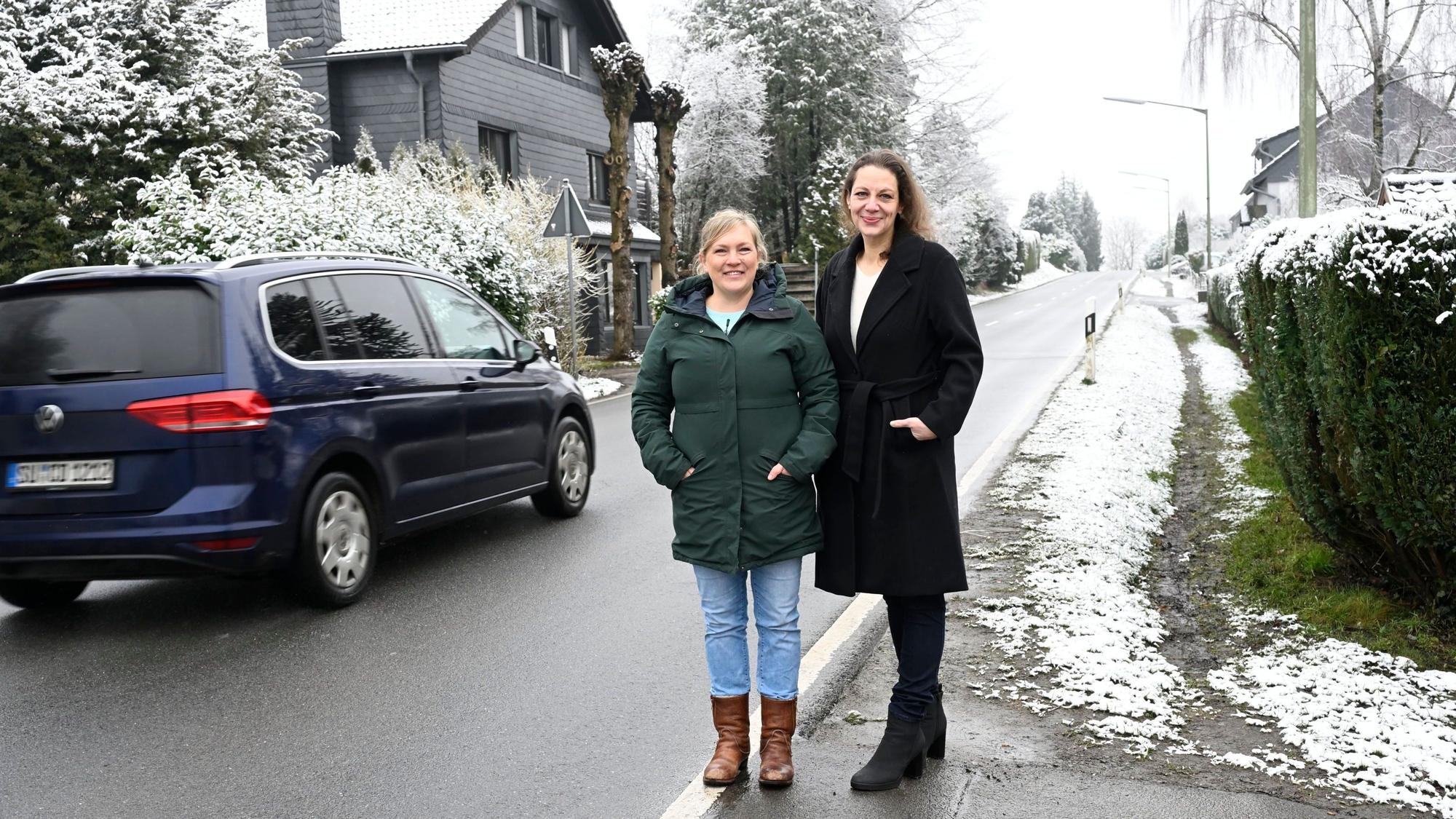 Zwei Frauen stehen neben einer Straße während ein Auto an ihnen vorbeifährt und lächeln in die Kamera.
