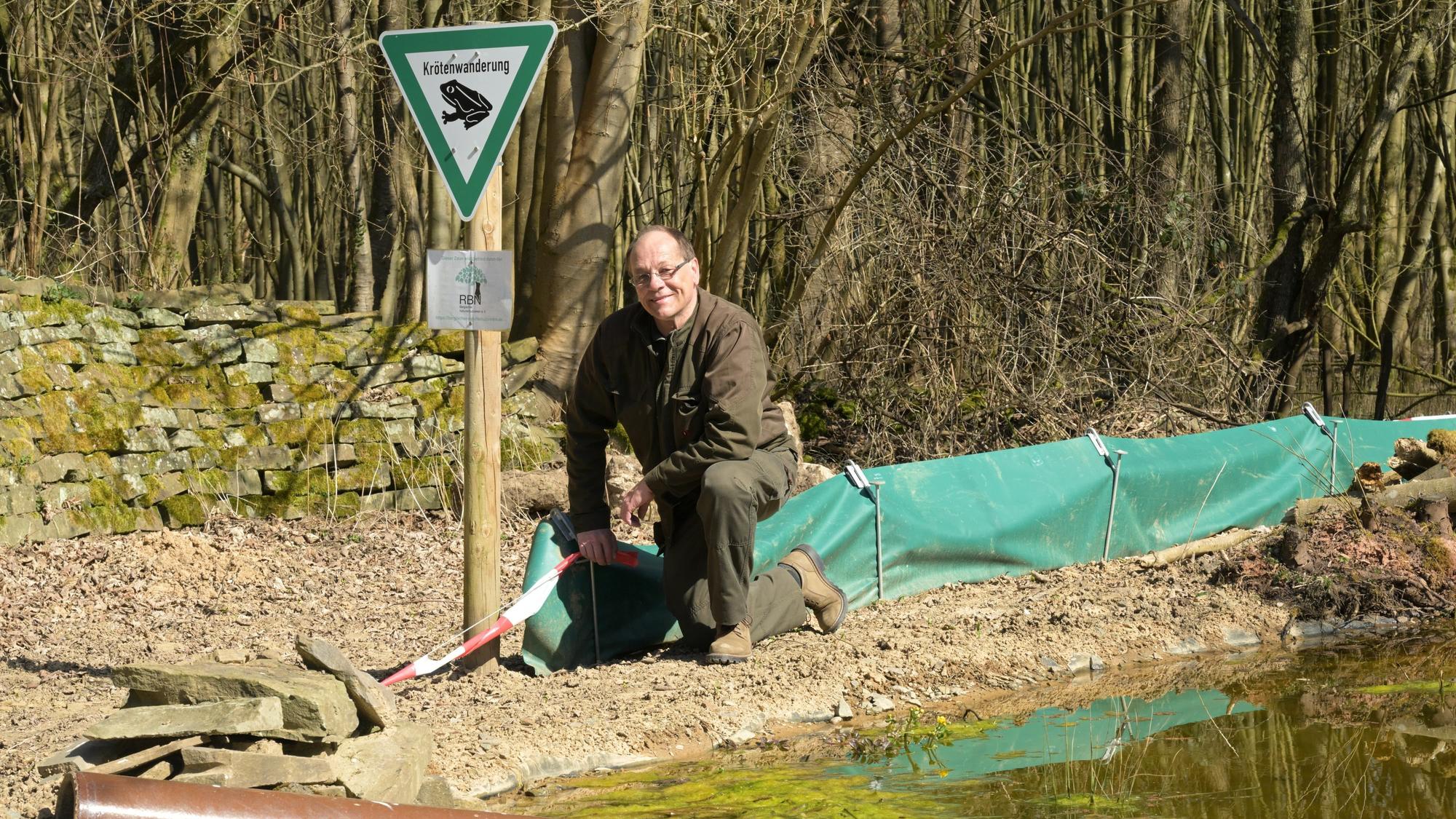 Ein Mann kniet an einem grünen Amphibienzaun an einem Gewässer.