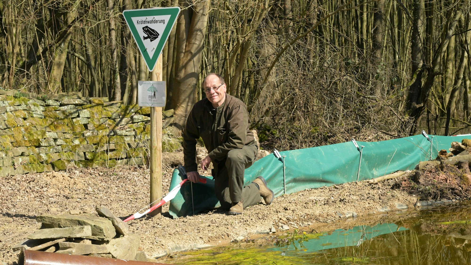 Ein Mann kniet an einem grünen Amphibienzaun an einem Gewässer.