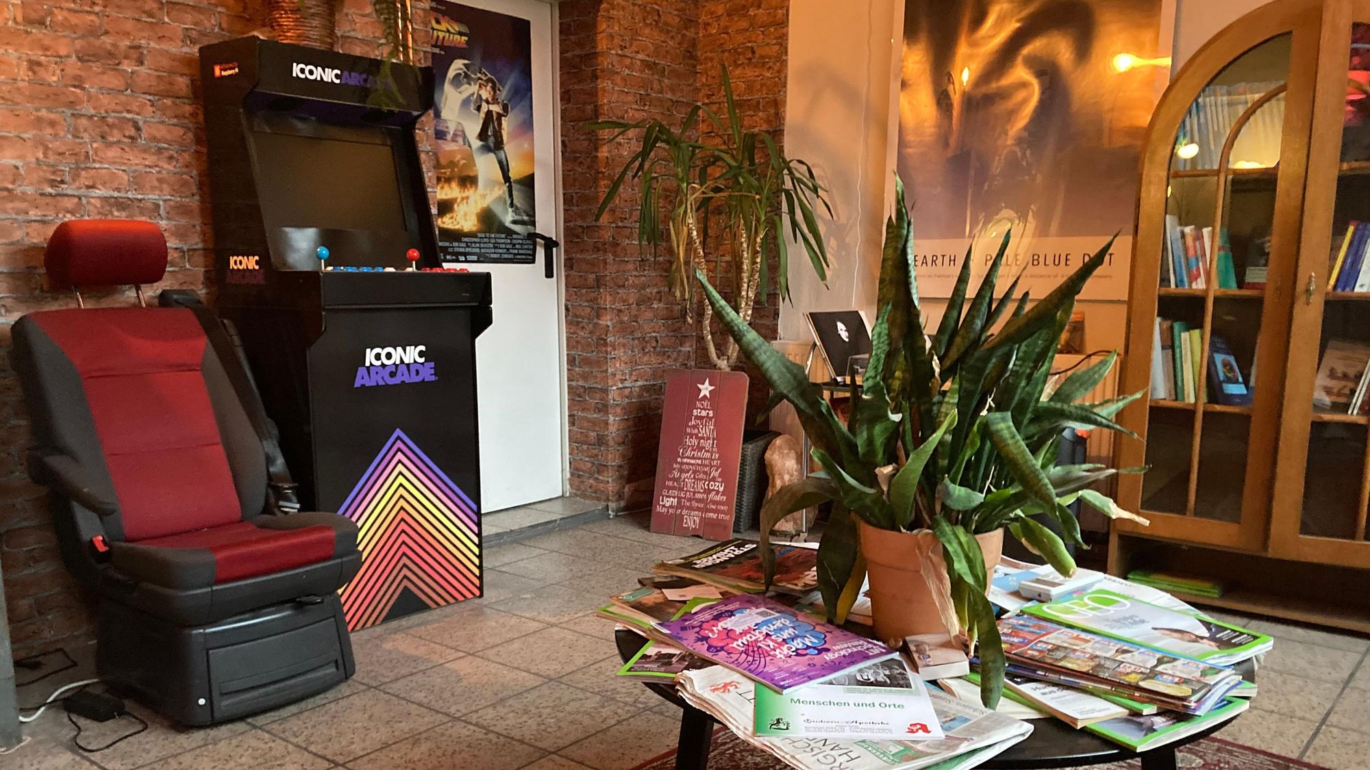Im Wartezimmer stehen ein Tisch mit Zeitschriften und einer Pflanze, ein Bücherregal und ein Arcade-Spielautomat.